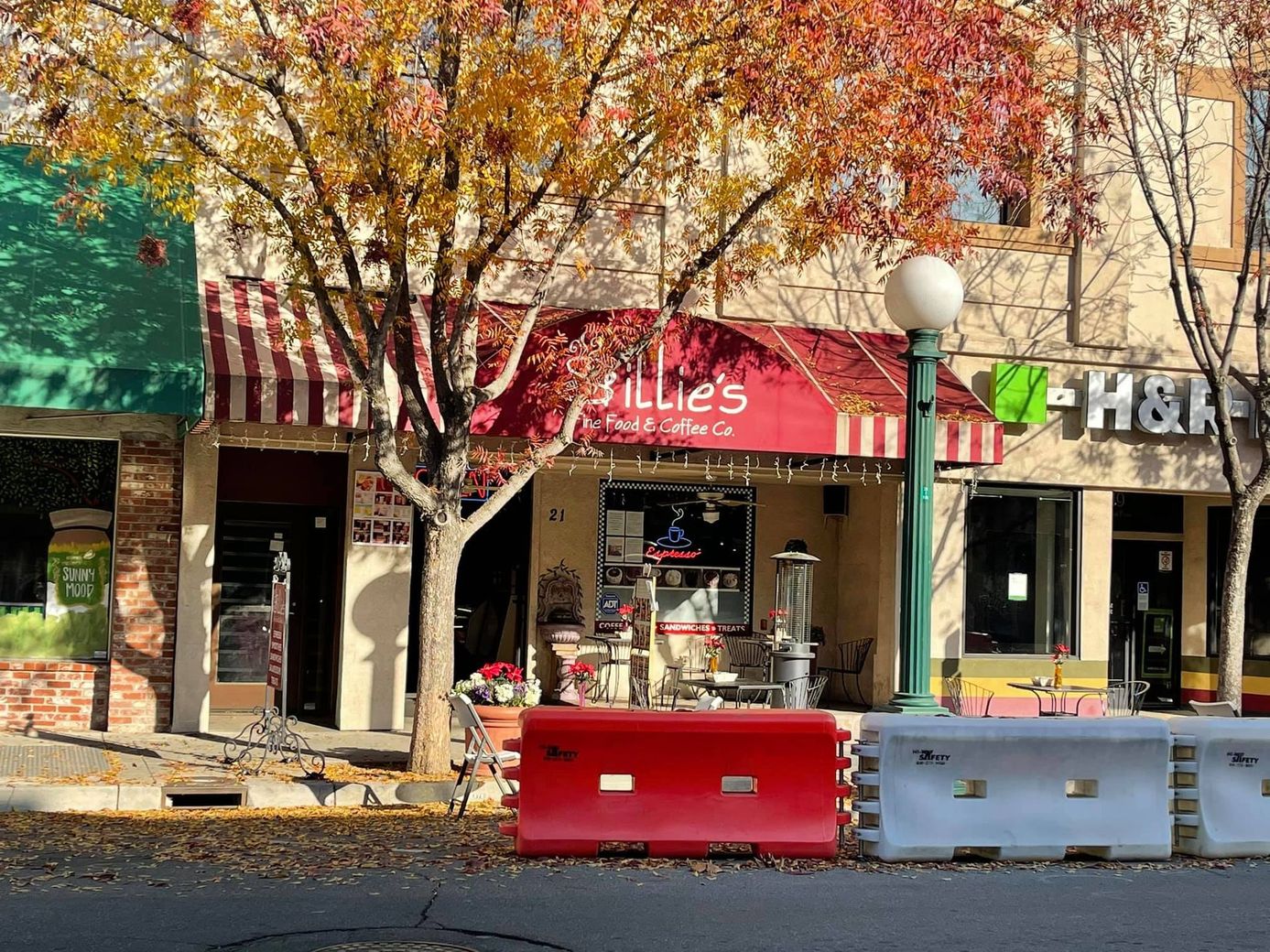 Downtown storefronts under autumn trees with red and white barricades in front