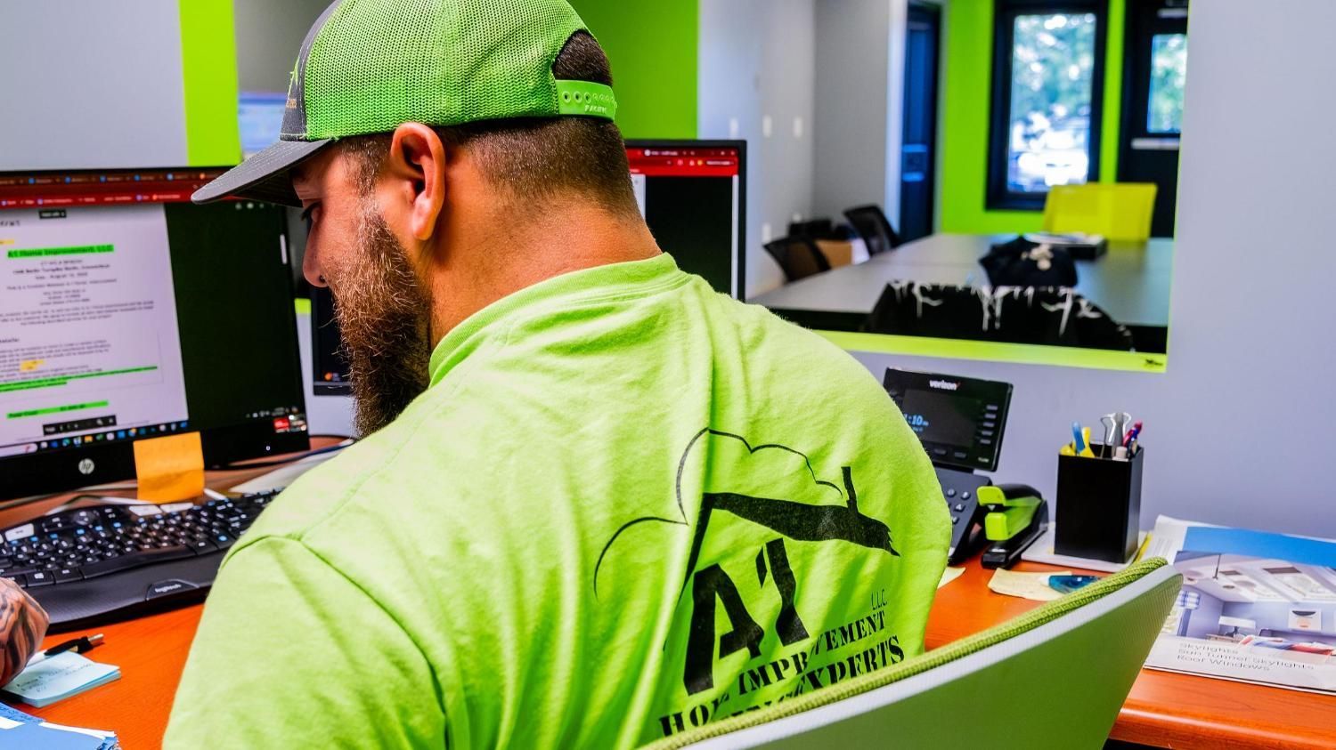A man wearing a green hat and a green shirt is sitting at a desk in front of a computer.