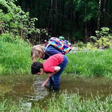 two children standing in a puddle, bent over to look at what's in it