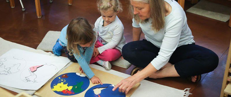 two preschool aged children sitting on the floor with a guide, working with the continents puzzle map