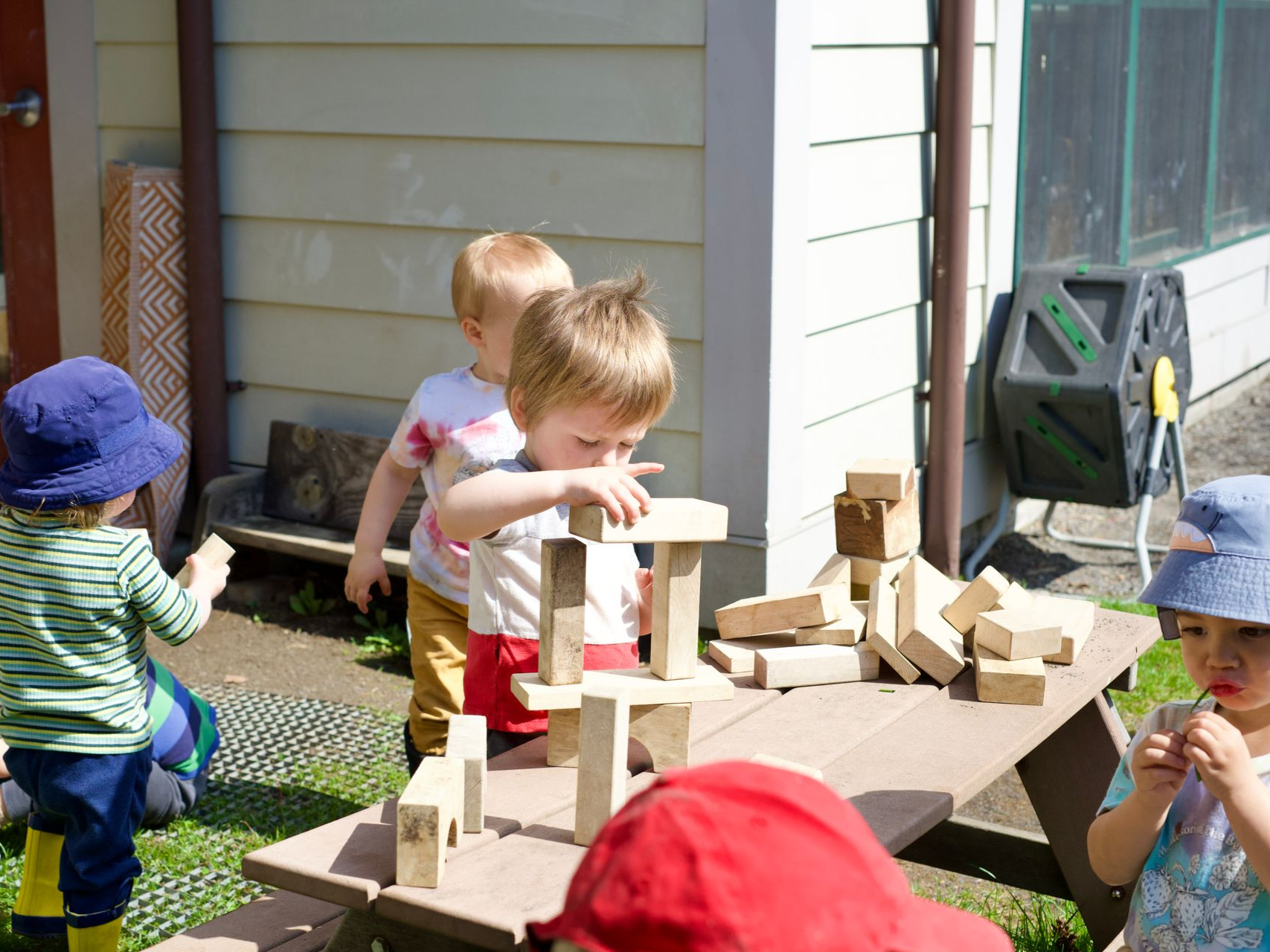 Group of toddlers exploring various materials in a grassy, sunny outdoor space