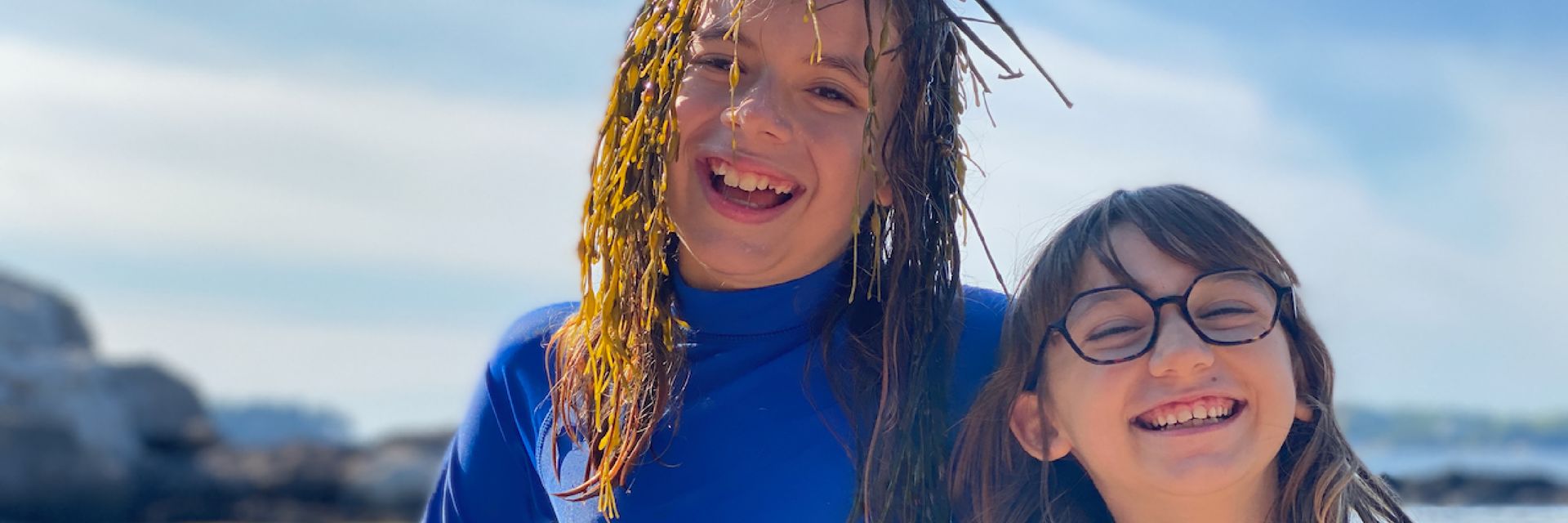 Image of two elementary aged children at the beach, side by side and smiling at the camera