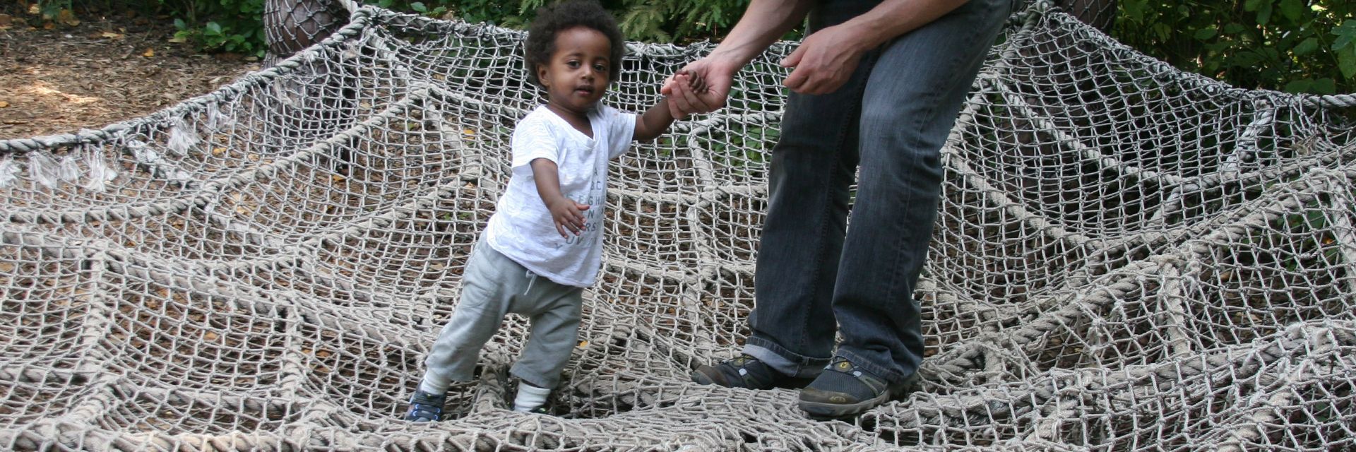 A toddler standing in a hanging rope web holding the hand of a nearby adult