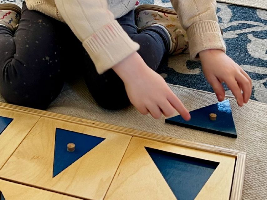 Image of a young child's legs and hands, sitting on the floor working with geometry materials.