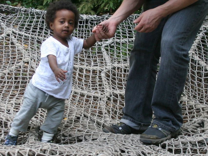A toddler standing in a hanging rope web holding the hand of a nearby adult