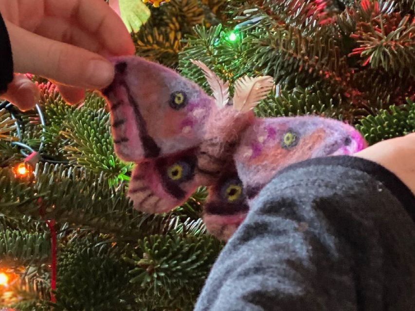 Image of a child's hand placing a felted moth ornament onto the branches of a Christmas tree