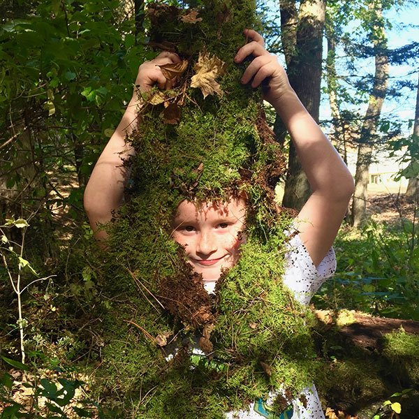 Child holding a large piece of moss covered bark in front of him, with his face peering through a hole in the bark