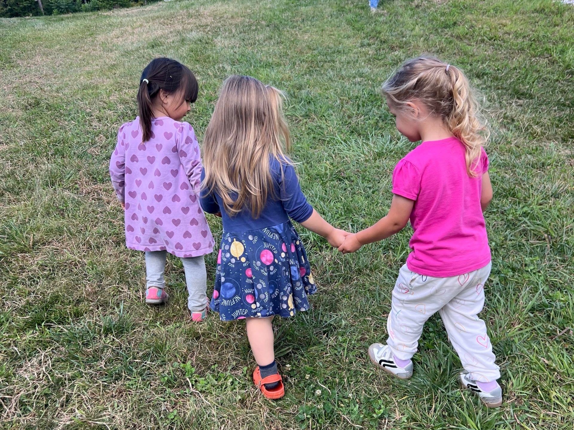 Image of three preschool girls in dresses holding hands, walking across grass