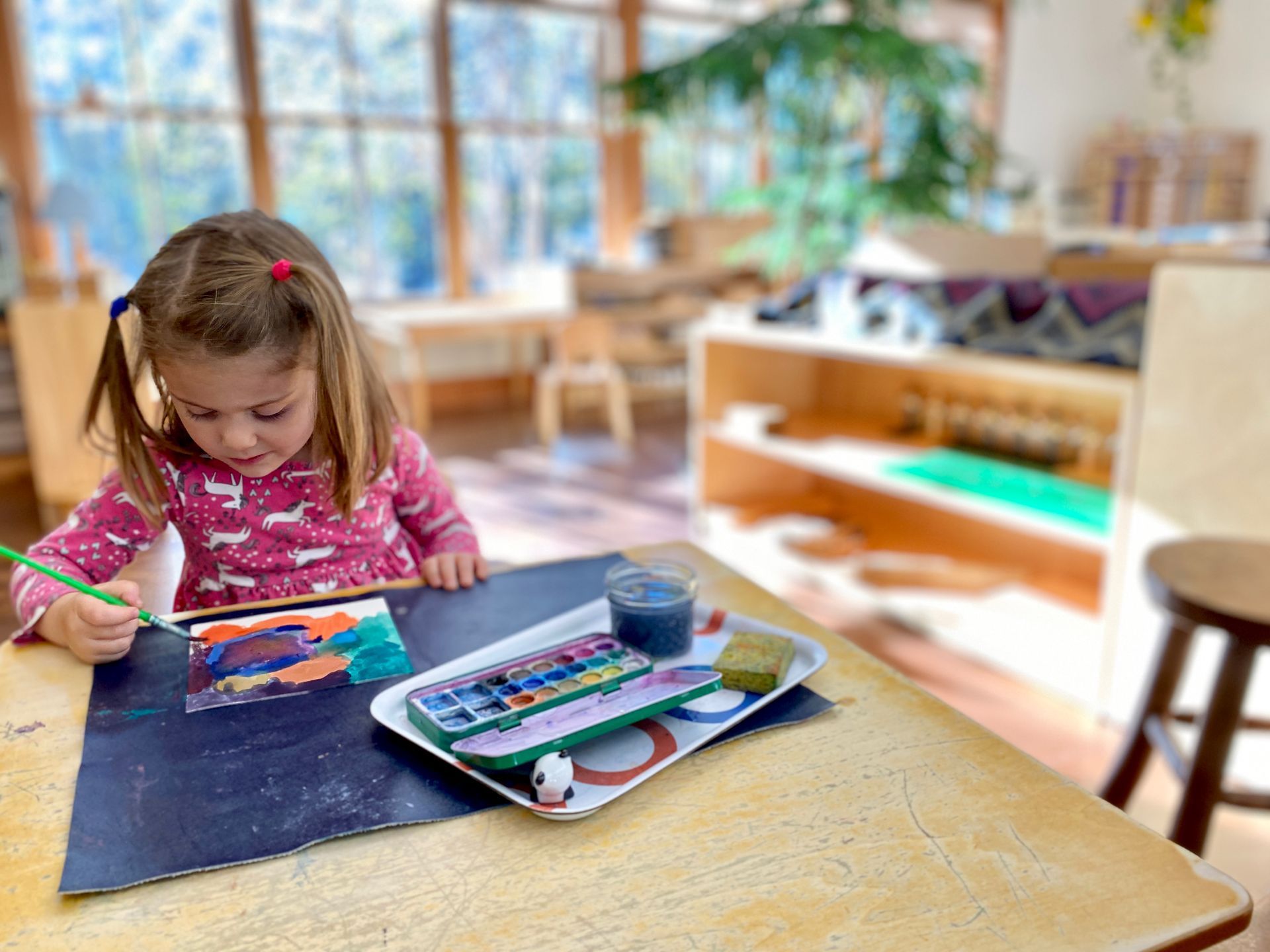 Image of a preschool aged girl sitting at a small table with a tray of painting supplies