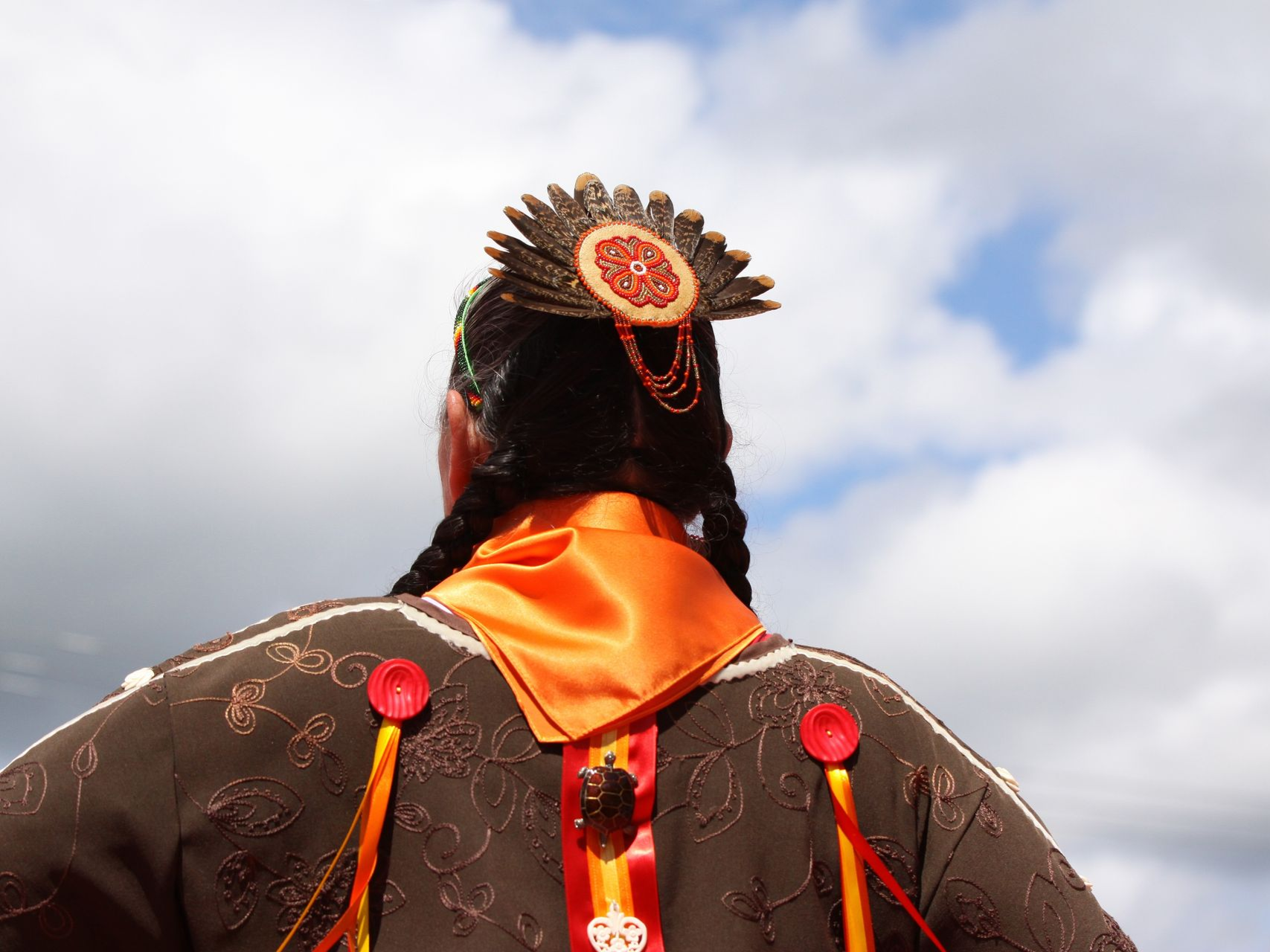 Image of head and shoulders of a person in indigenous dress with a blue cloudy sky in the background