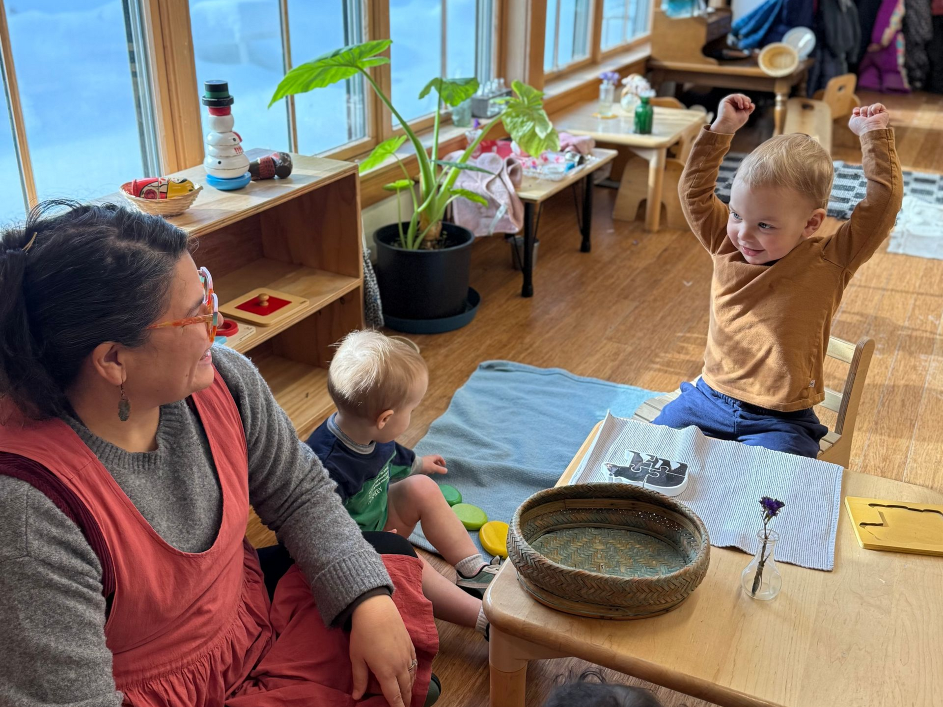Image of a toddler sitting at a table with arms raised in celebration while a smiling adult looks on
