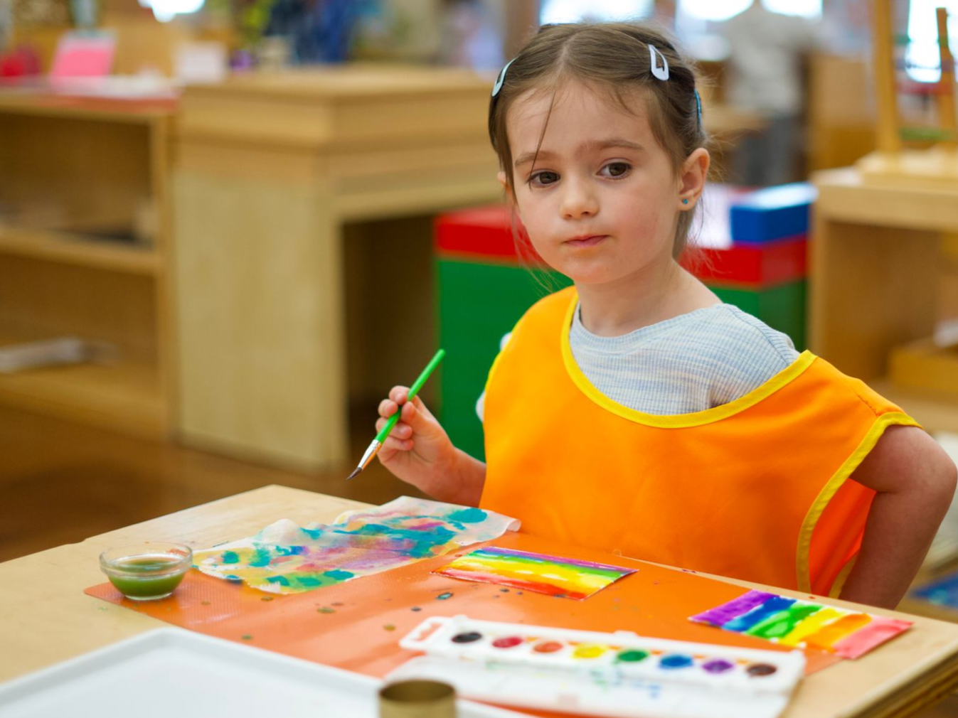 Image of a preschool aged girl sitting at a small table with a palette of water colors and paper