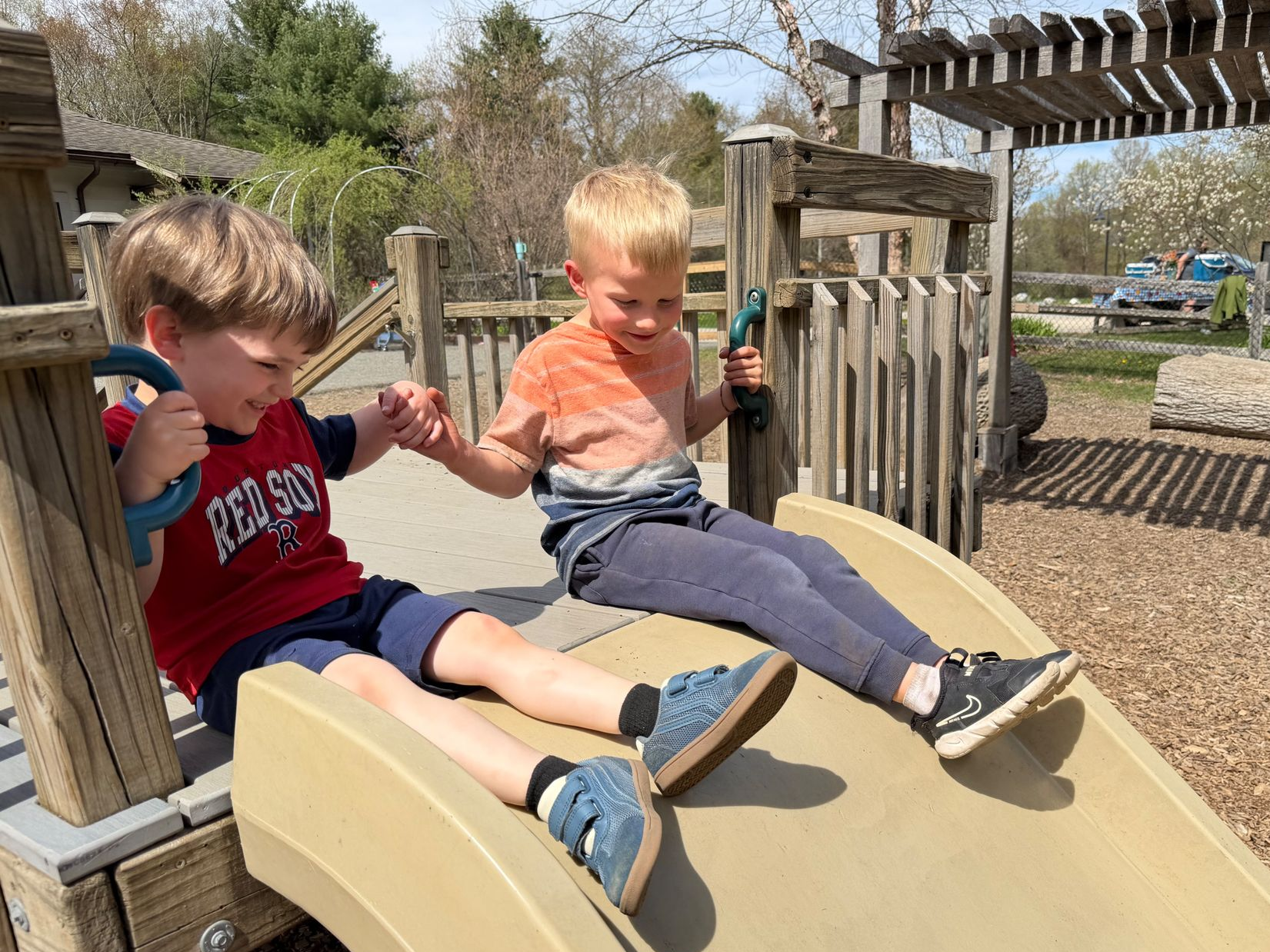 Two preschool aged children sitting at the top of a slide, smiling and holding hands