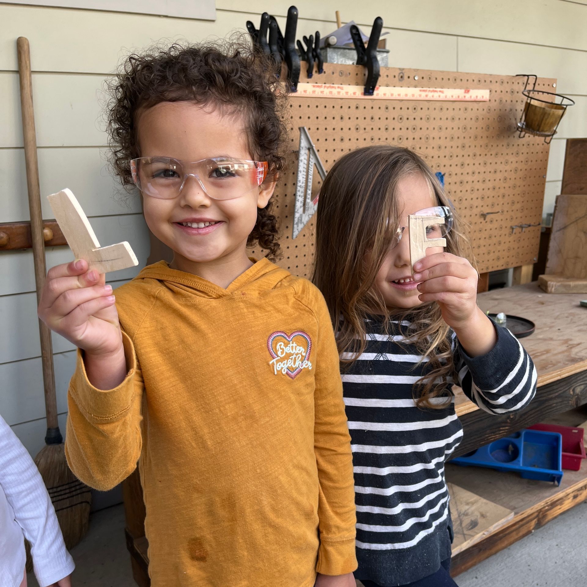 two preschool children in safety goggles holding wooden letters they cut out with a saw