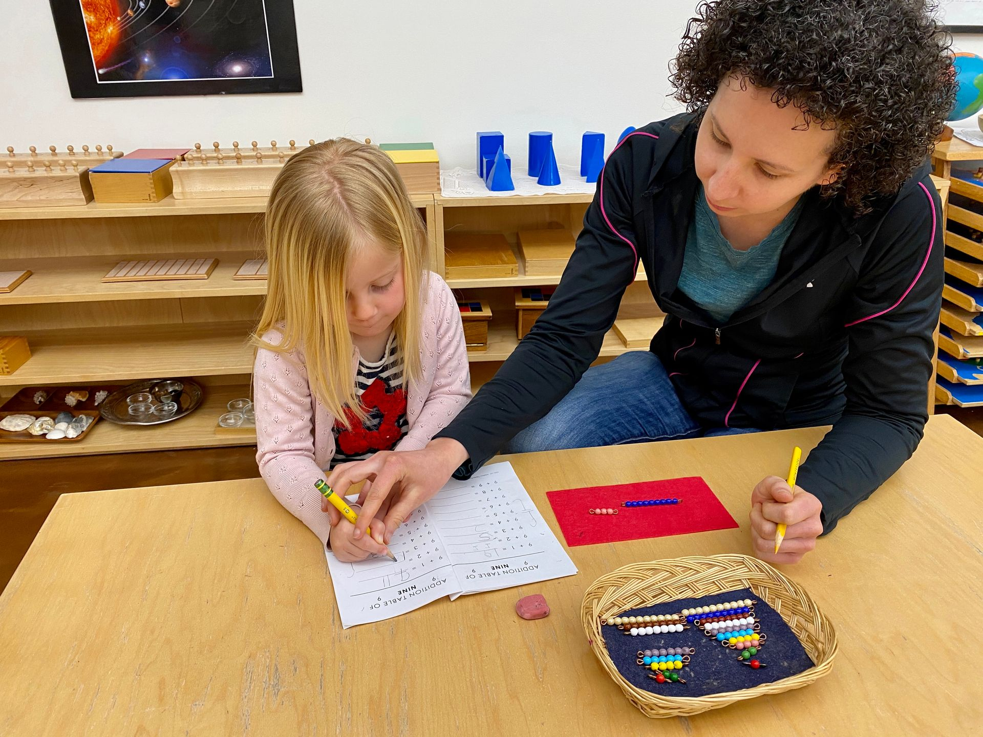 image of a preschool aged child sitting at a low table with a montessori guide helping with pencil g
