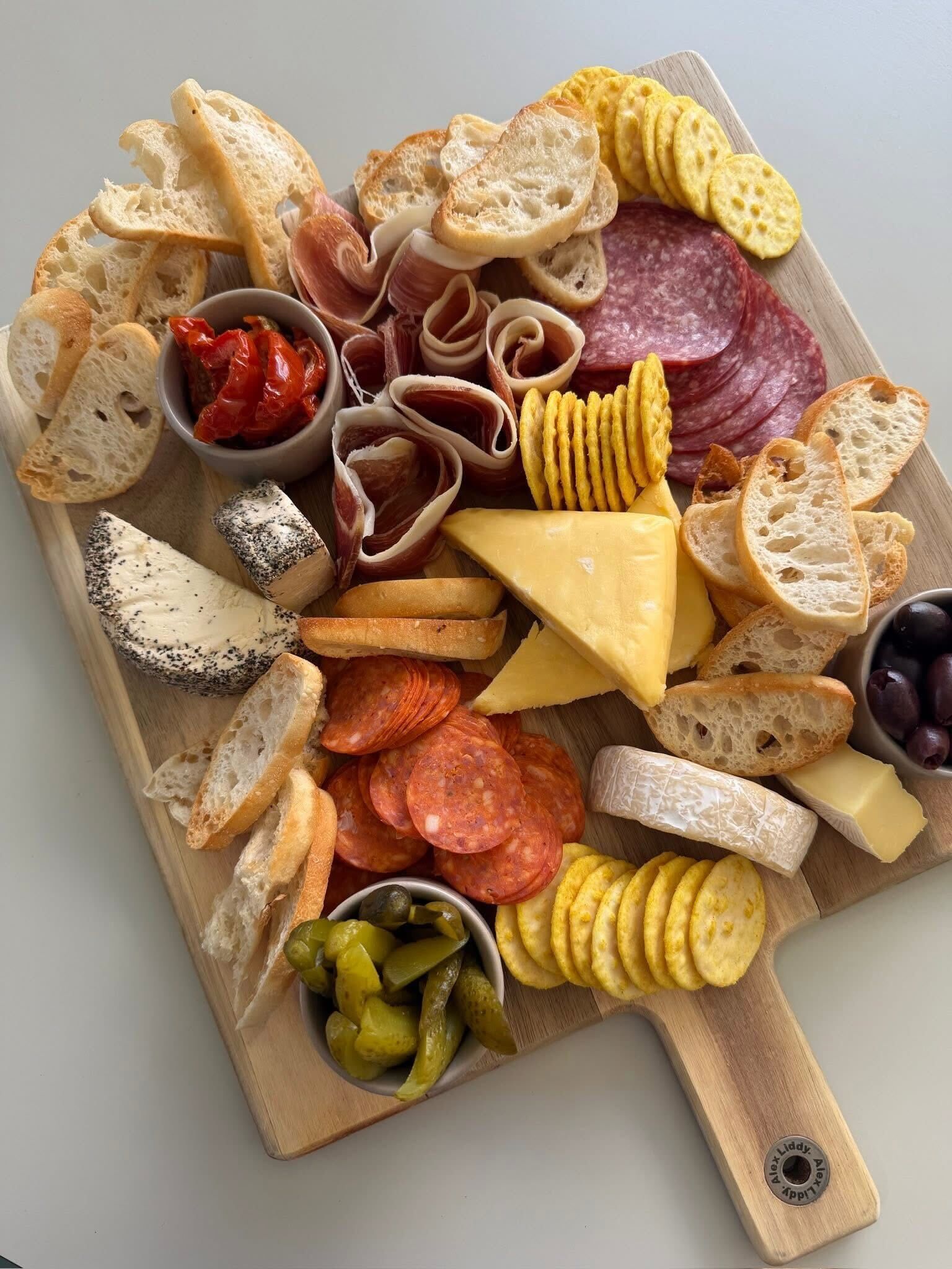 Charcuterie board: meats, cheeses, crackers, bread, olives, peppers, in bowls, on wooden cutting board.
