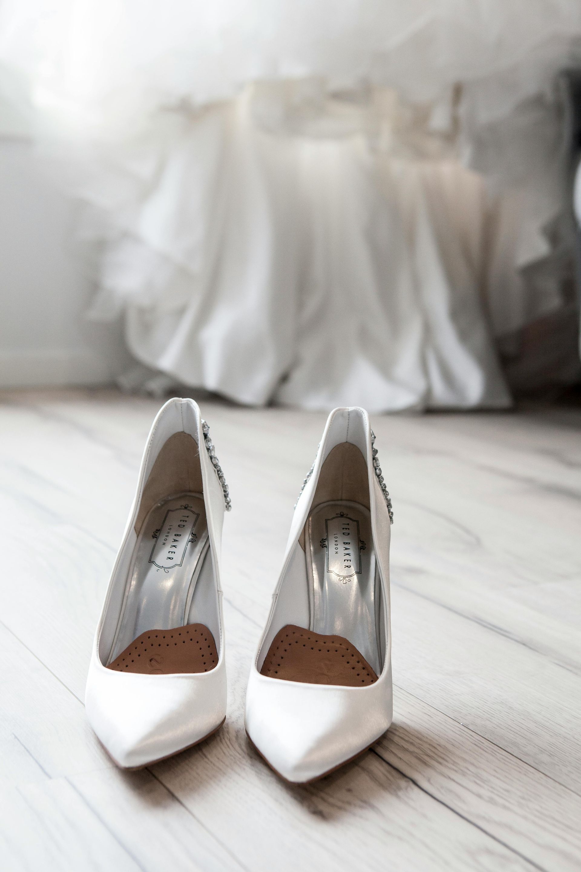 White bridal heels on a wooden floor, with a white wedding dress in the blurred background.