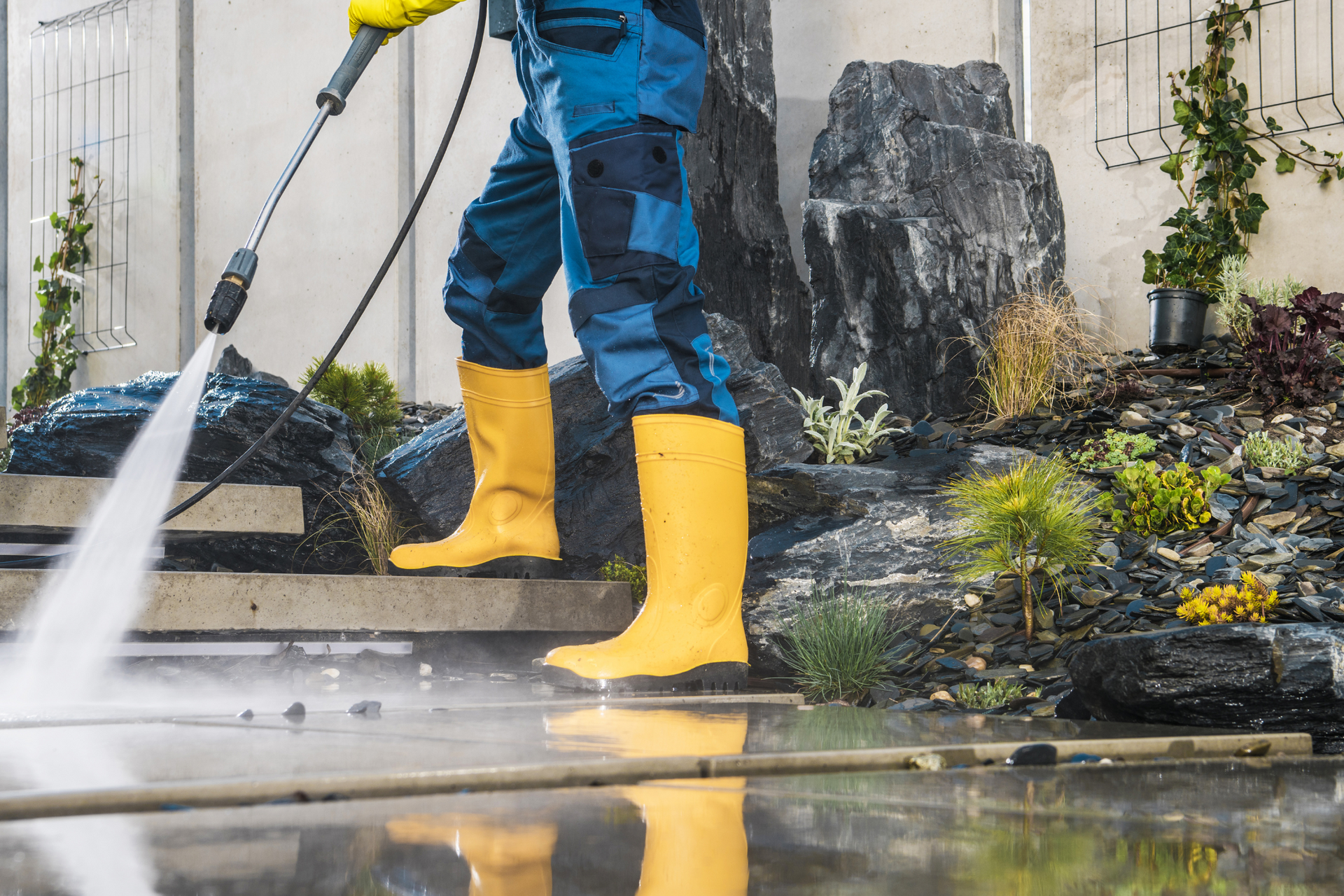 A man in yellow boots is using a high pressure washer to clean a sidewalk.