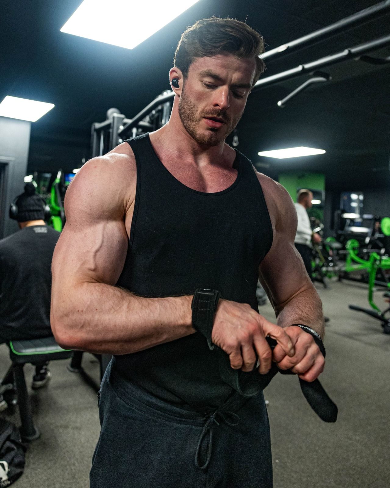 Muscular man in black tank top adjusting wrist straps in a gym with bright overhead lights