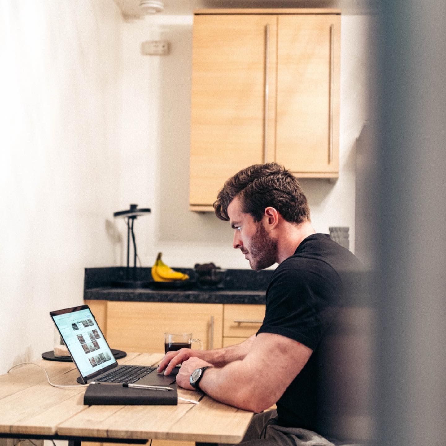 Man working on a laptop at a kitchen table with a black keyboard and banana on the counter behind him