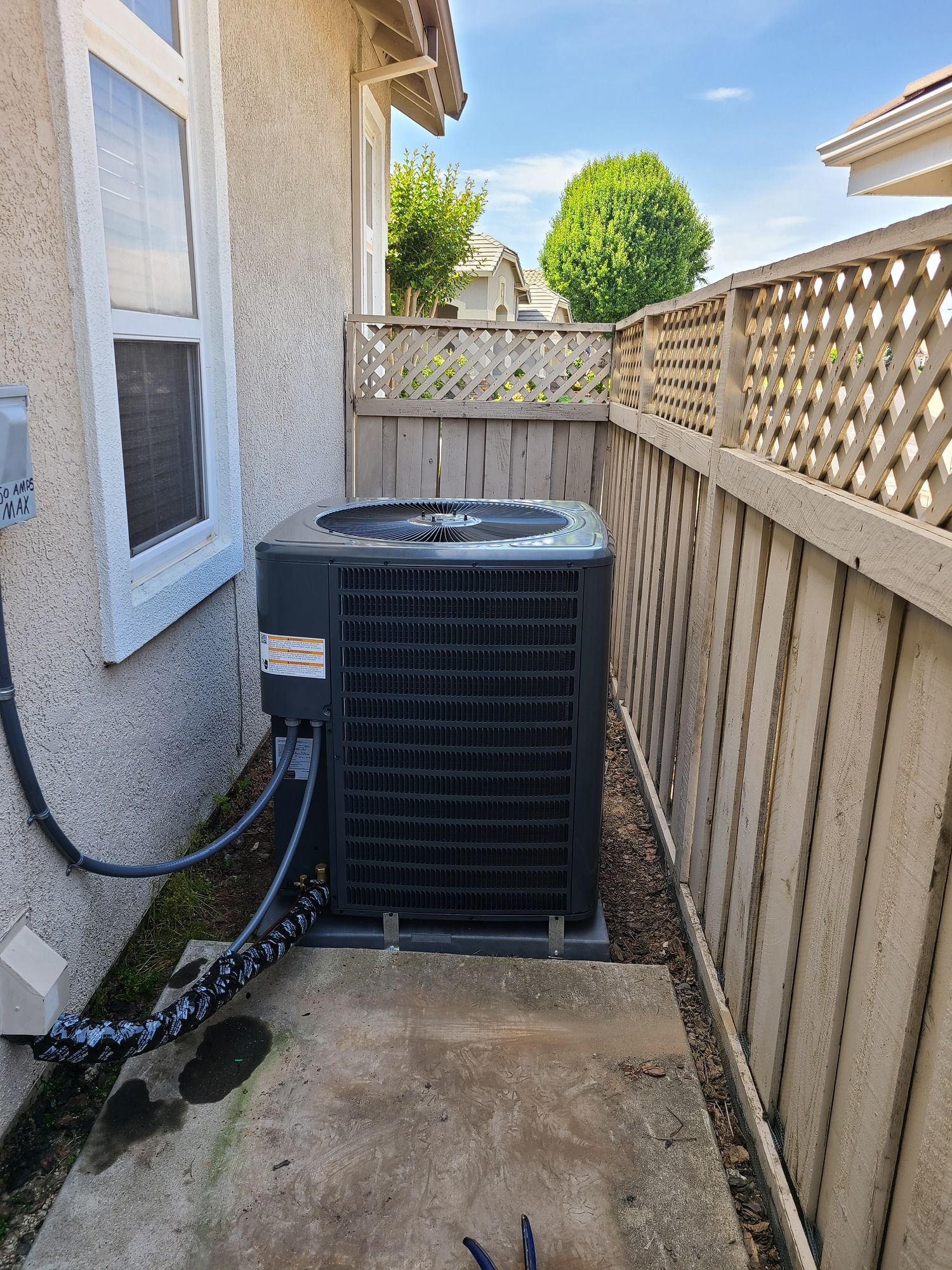 An air conditioning unit is against a wall and fence, outdoors on a patio.