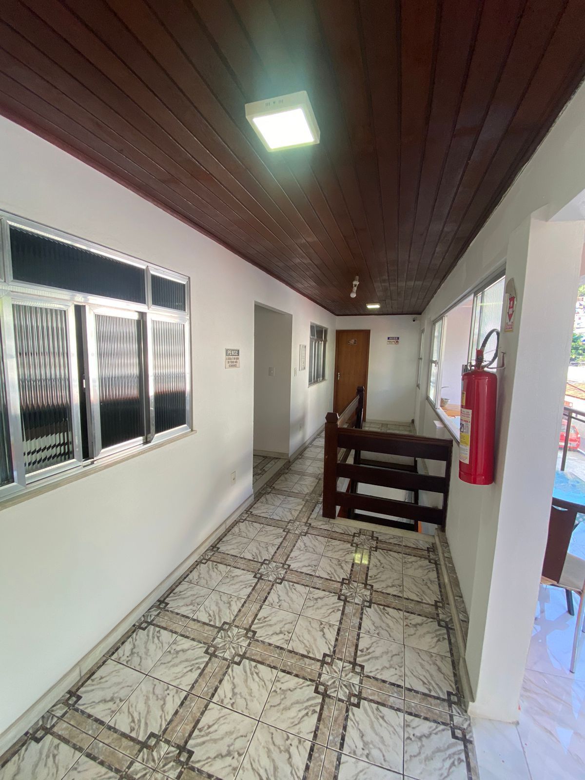 Corredor comprido com piso de azulejo, janelas, teto de madeira e extintor de incêndio.