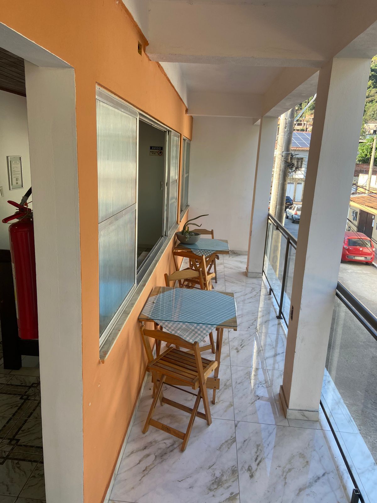 Varanda com mesinhas e cadeiras; parede laranja com janelas; a rua é visível do lado de fora da grade.