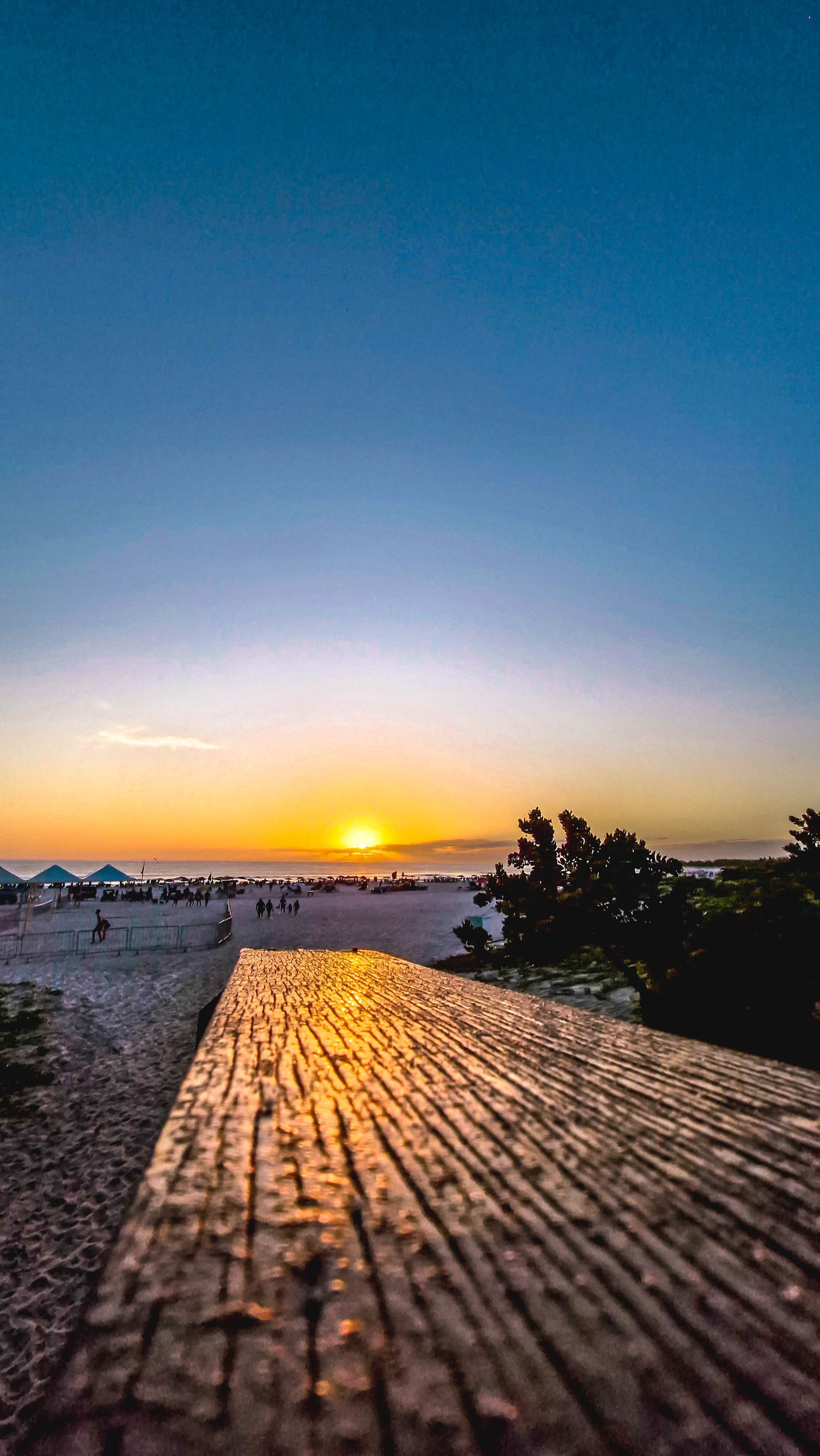 Pôr do sol na praia, com passarela de madeira em primeiro plano. Sol alaranjado, céu azul, pessoas na areia.