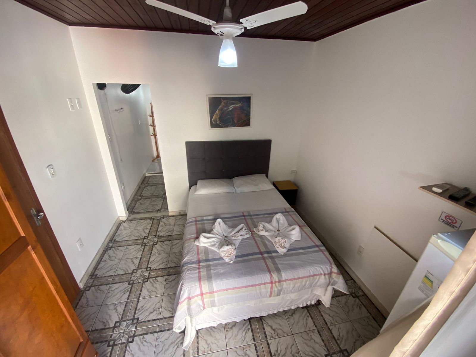 Quarto de hotel pequeno com cama, mesa de cabeceira e frigobar. Toalhas em formato de cisne sobre a cama.