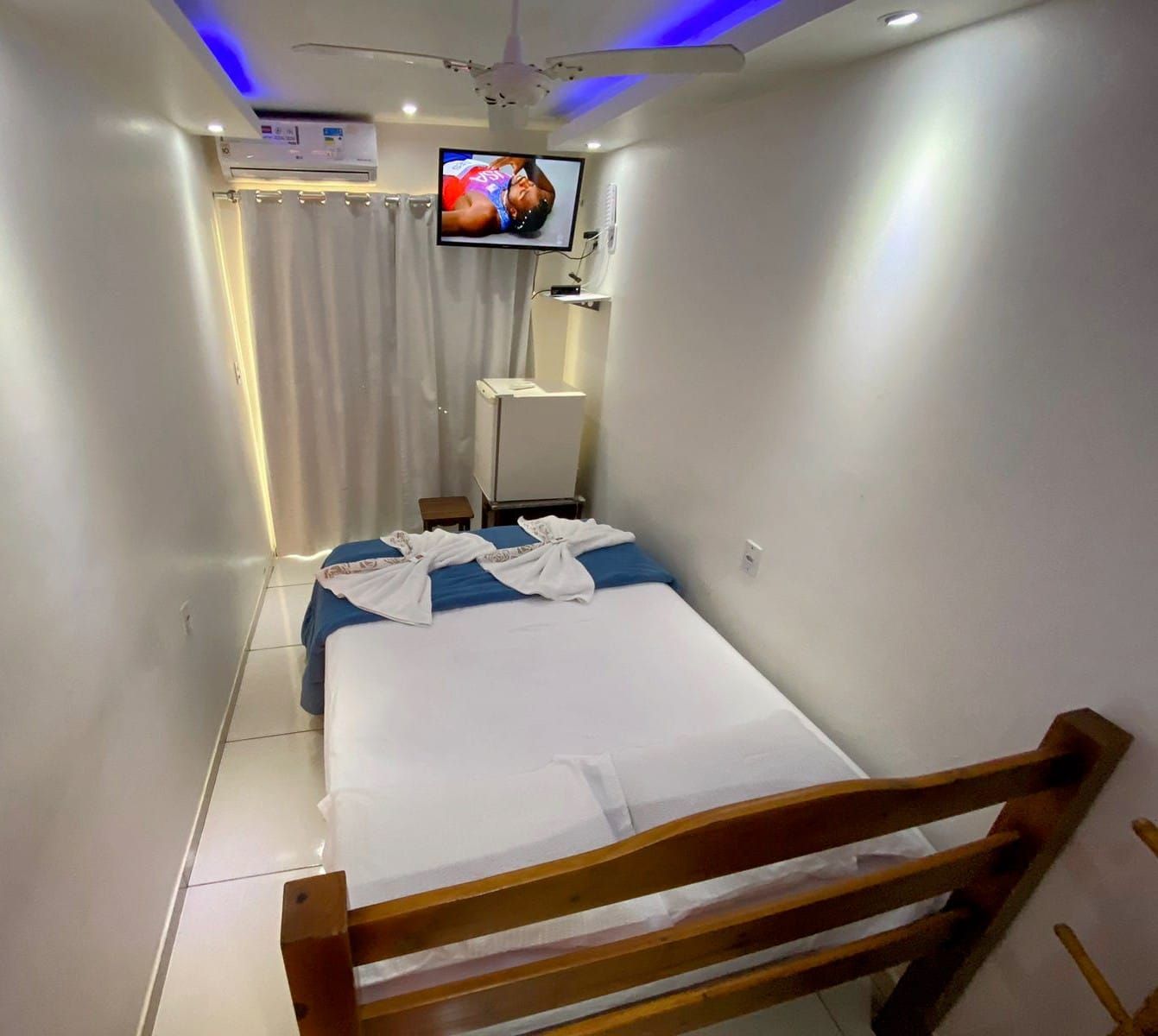 Quarto de hotel pequeno com cama, TV e frigobar. Paredes brancas com detalhes em azul.