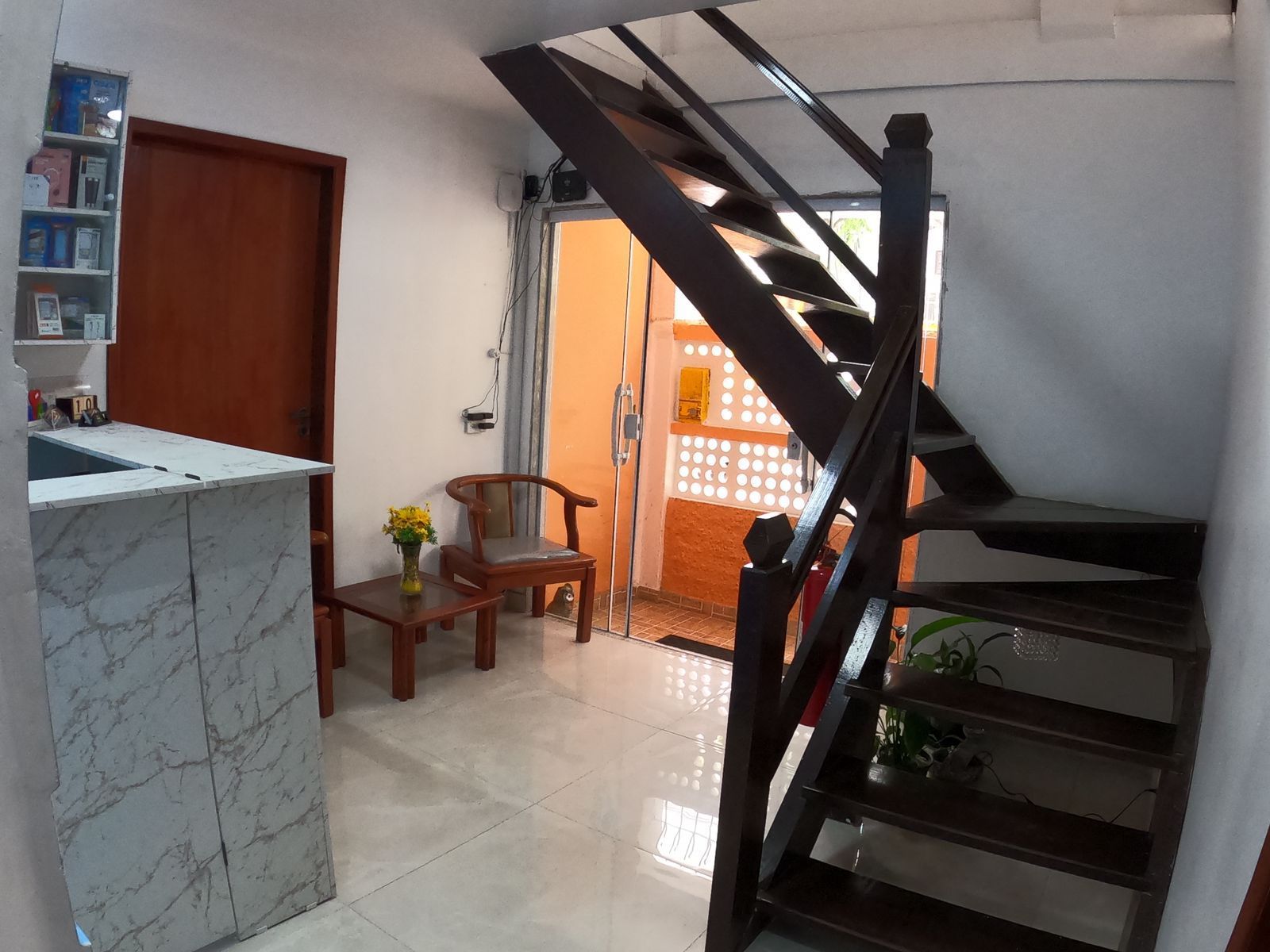 Interior com escada de madeira, balcão de mármore, porta, mesinha com cadeira e pequena estante.