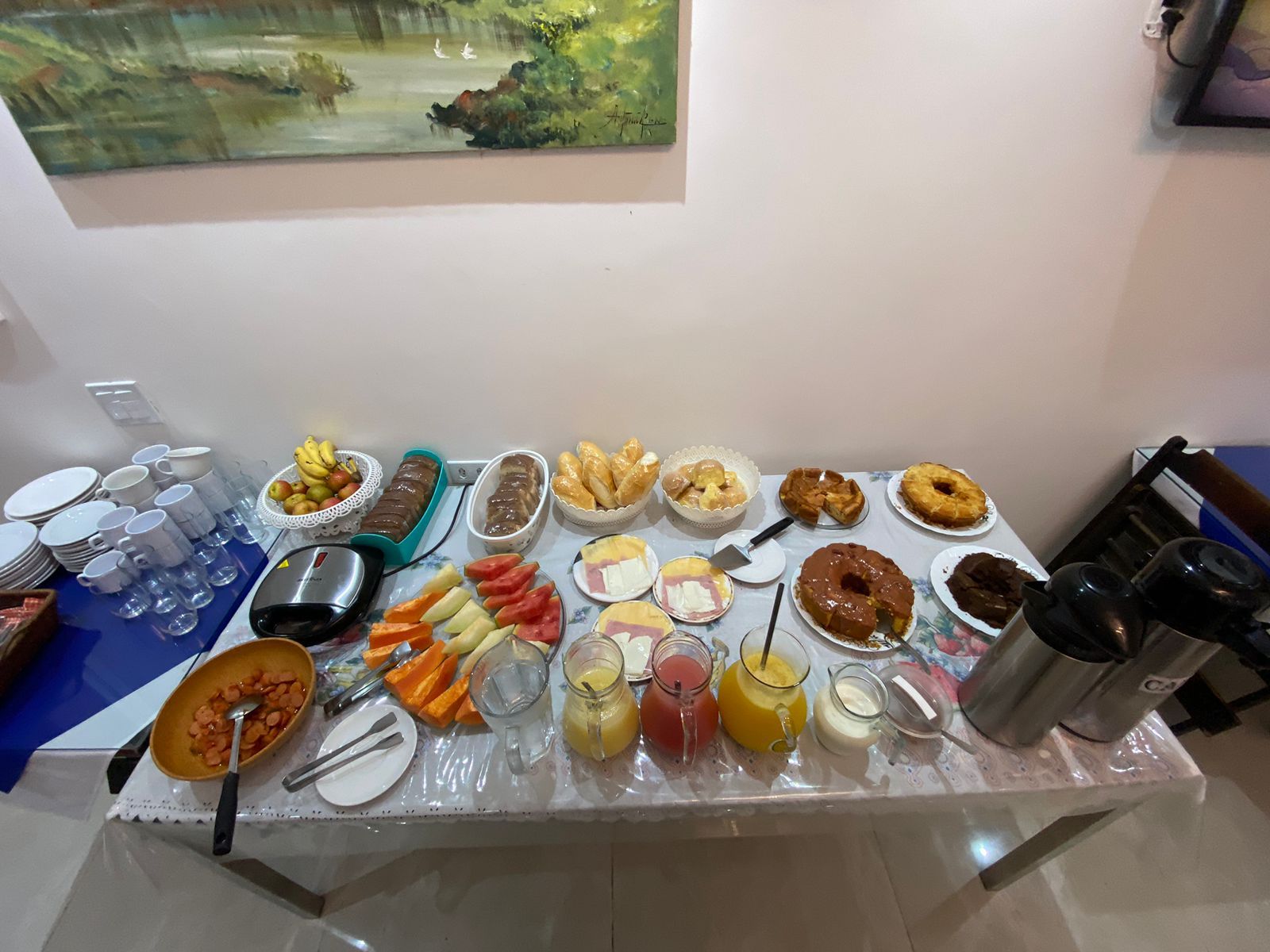 Buffet de café da manhã com frutas, doces e bebidas em uma mesa encostada em uma parede com obras de arte.