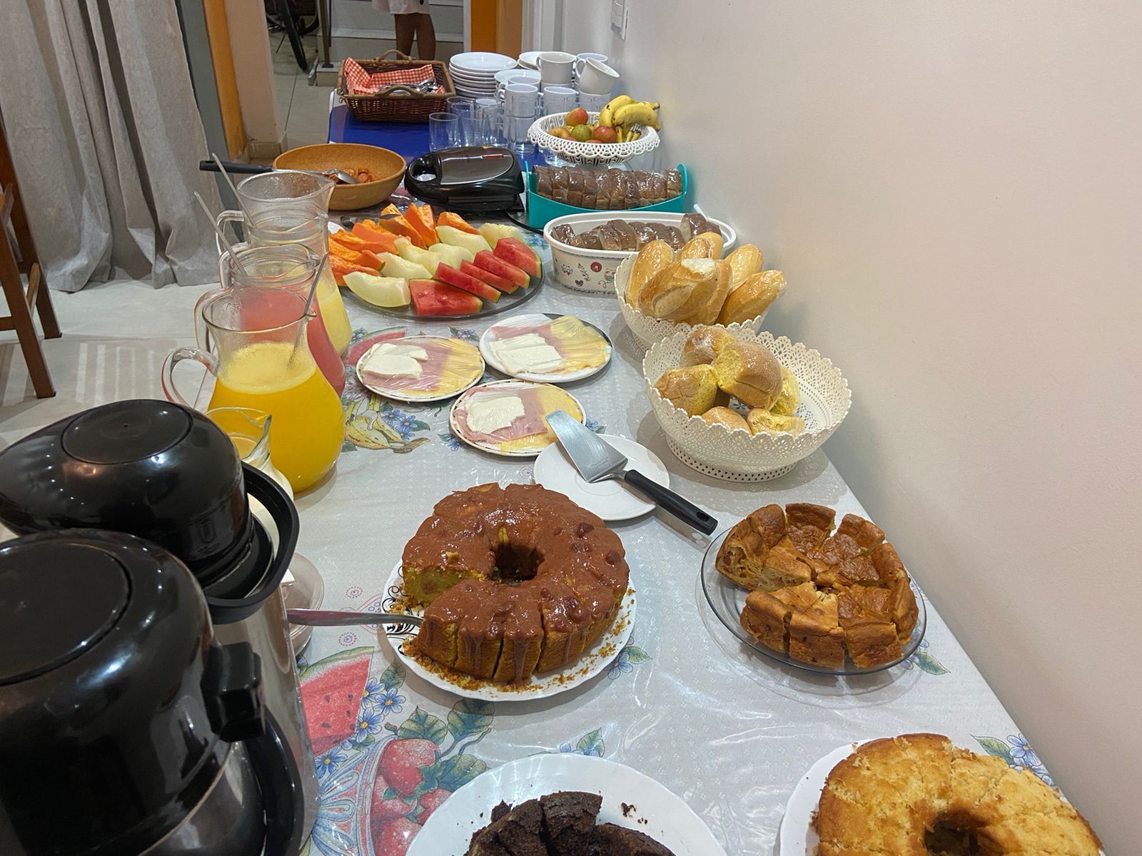 Mesa de buffet de café da manhã com bolos, frutas, pães e bebidas.