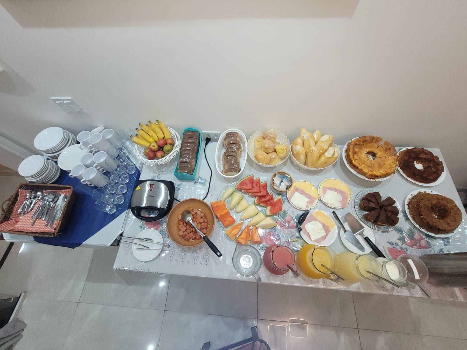 Buffet de café da manhã com frutas, bolos, doces, café e copos dispostos em uma mesa.