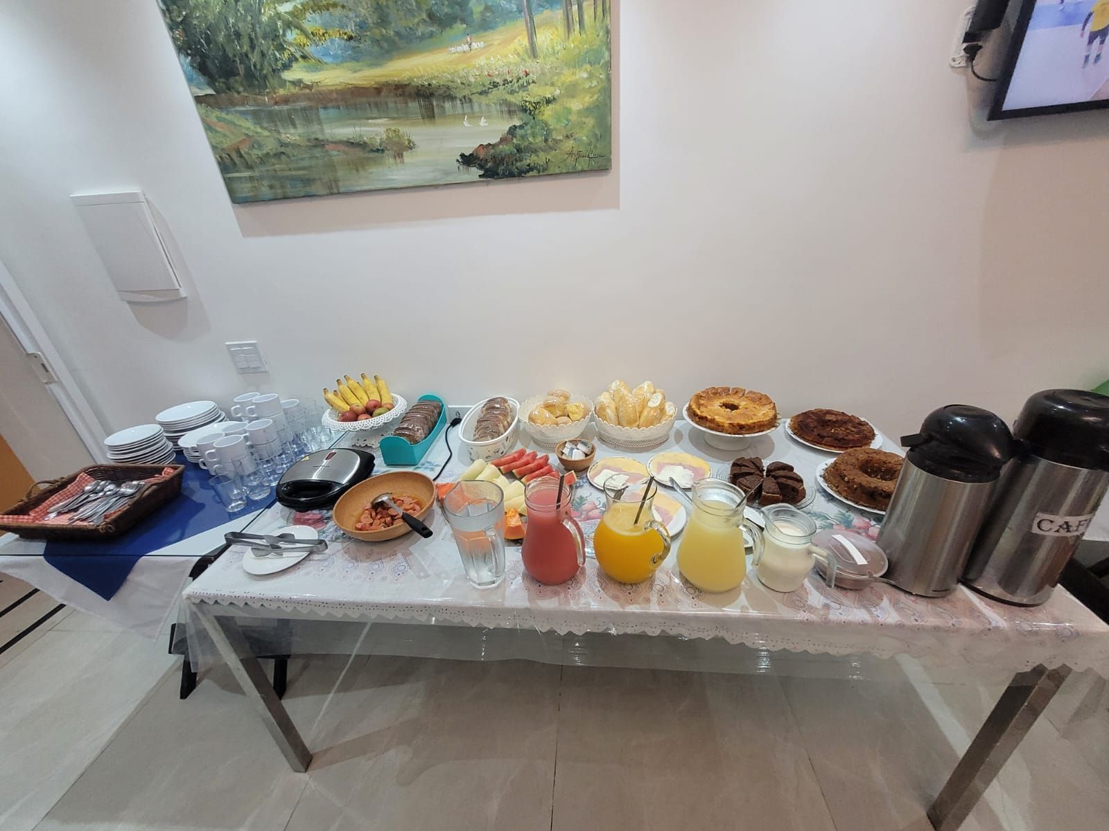 Mesa repleta de itens para o café da manhã: frutas, doces, suco e garrafas térmicas de café.