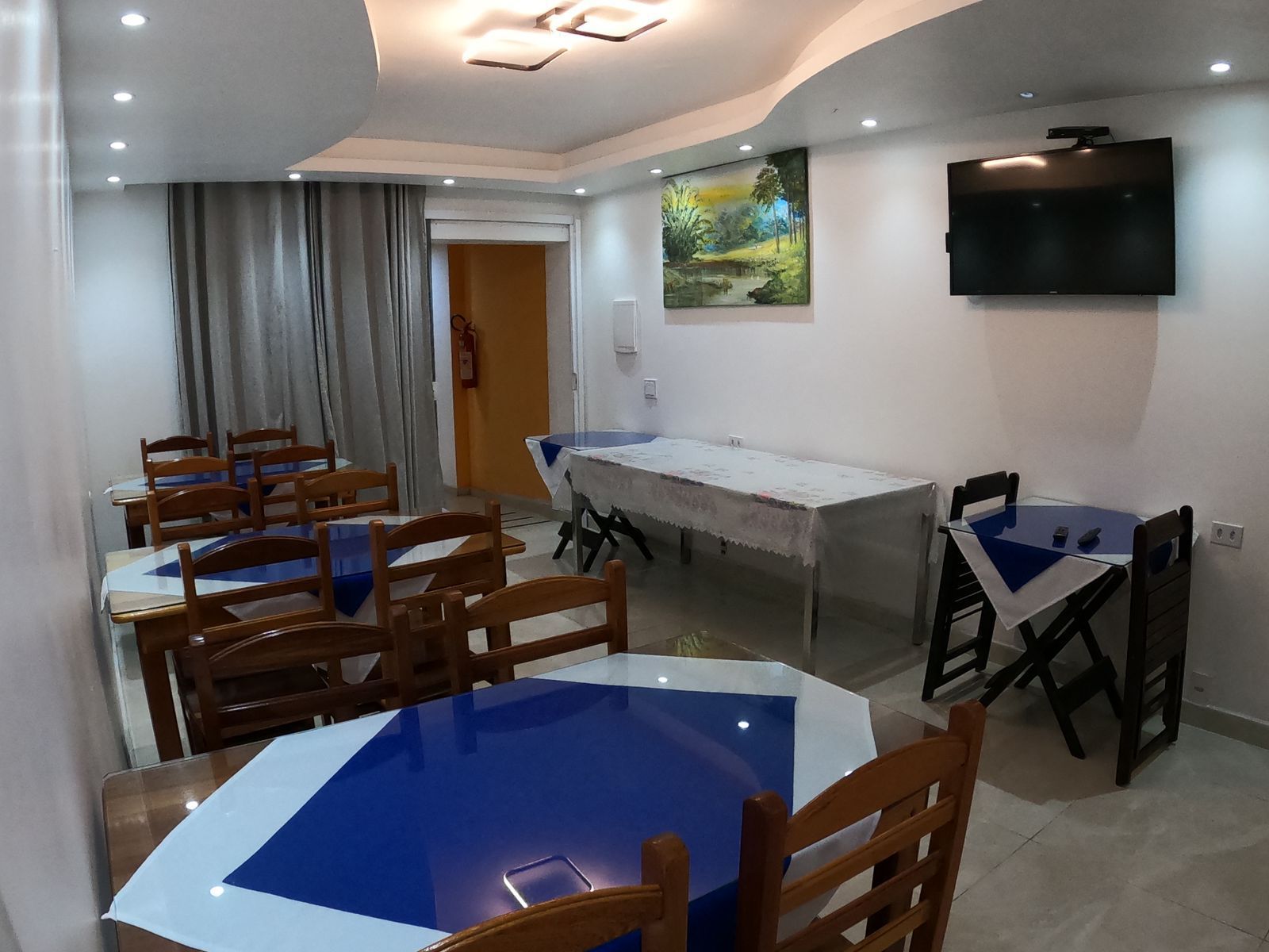 Sala de jantar com mesas e cadeiras, cobertas com toalhas azuis e brancas. Uma TV e um quadro estão na parede.