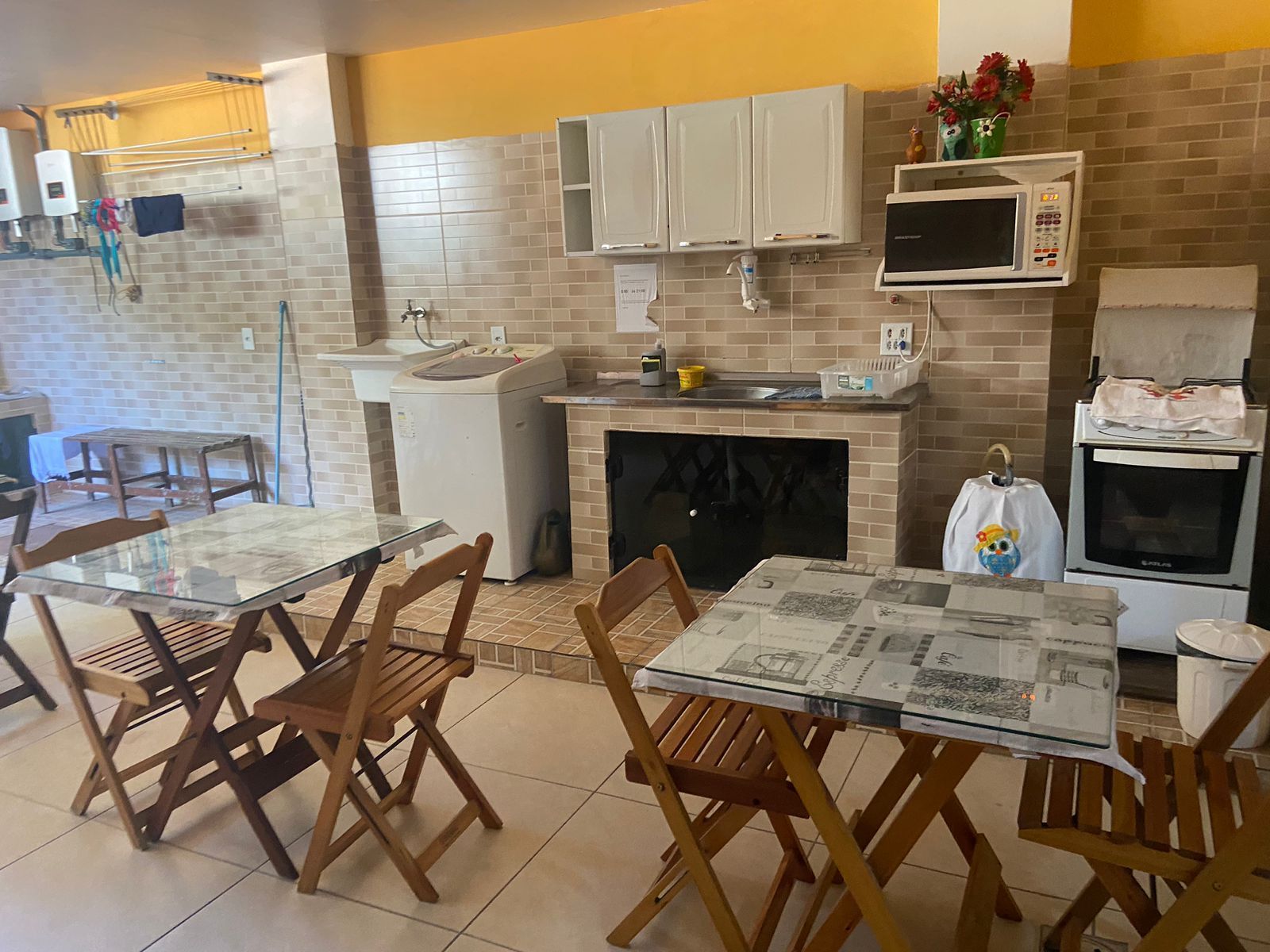 Cozinha com mesas e cadeiras, micro-ondas, armários e eletrodomésticos encostada em uma parede de tijolos.