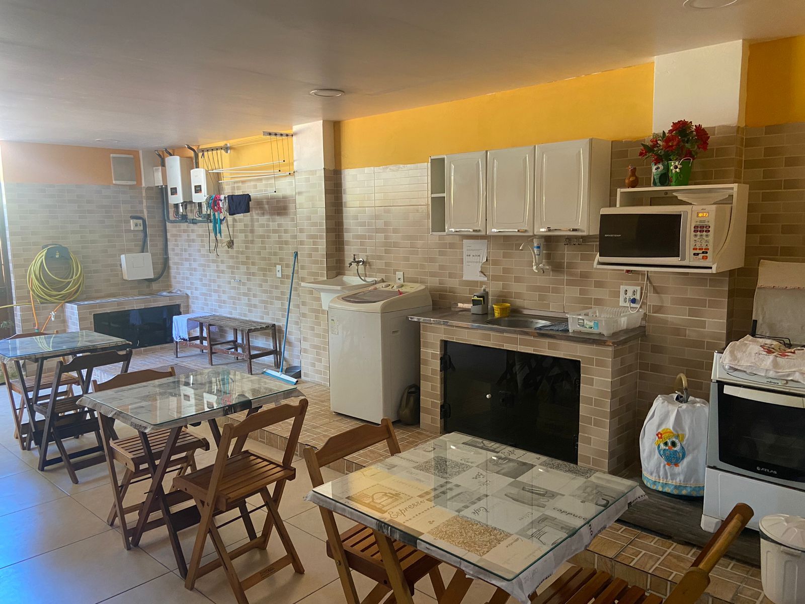 Cozinha pequena com mesas, armários, eletrodomésticos e micro-ondas. Paredes amarelas e bege.