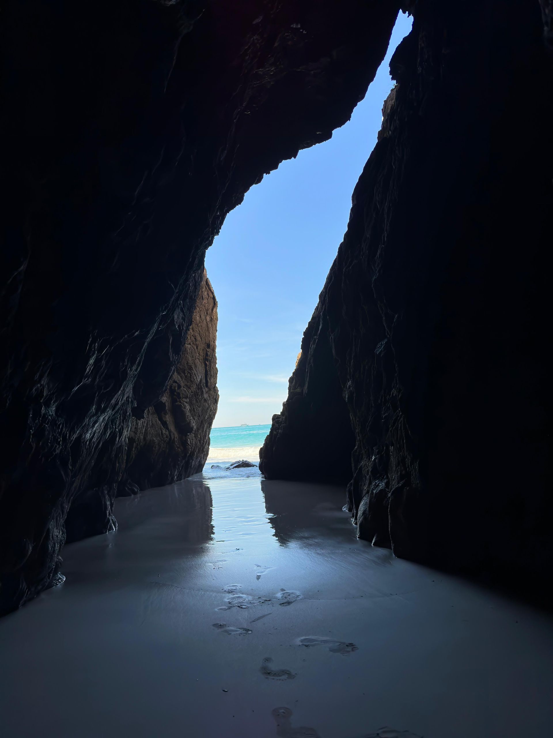 Entrada escura de uma caverna emoldurando a vista de uma praia de areia branca, água turquesa e céu azul.