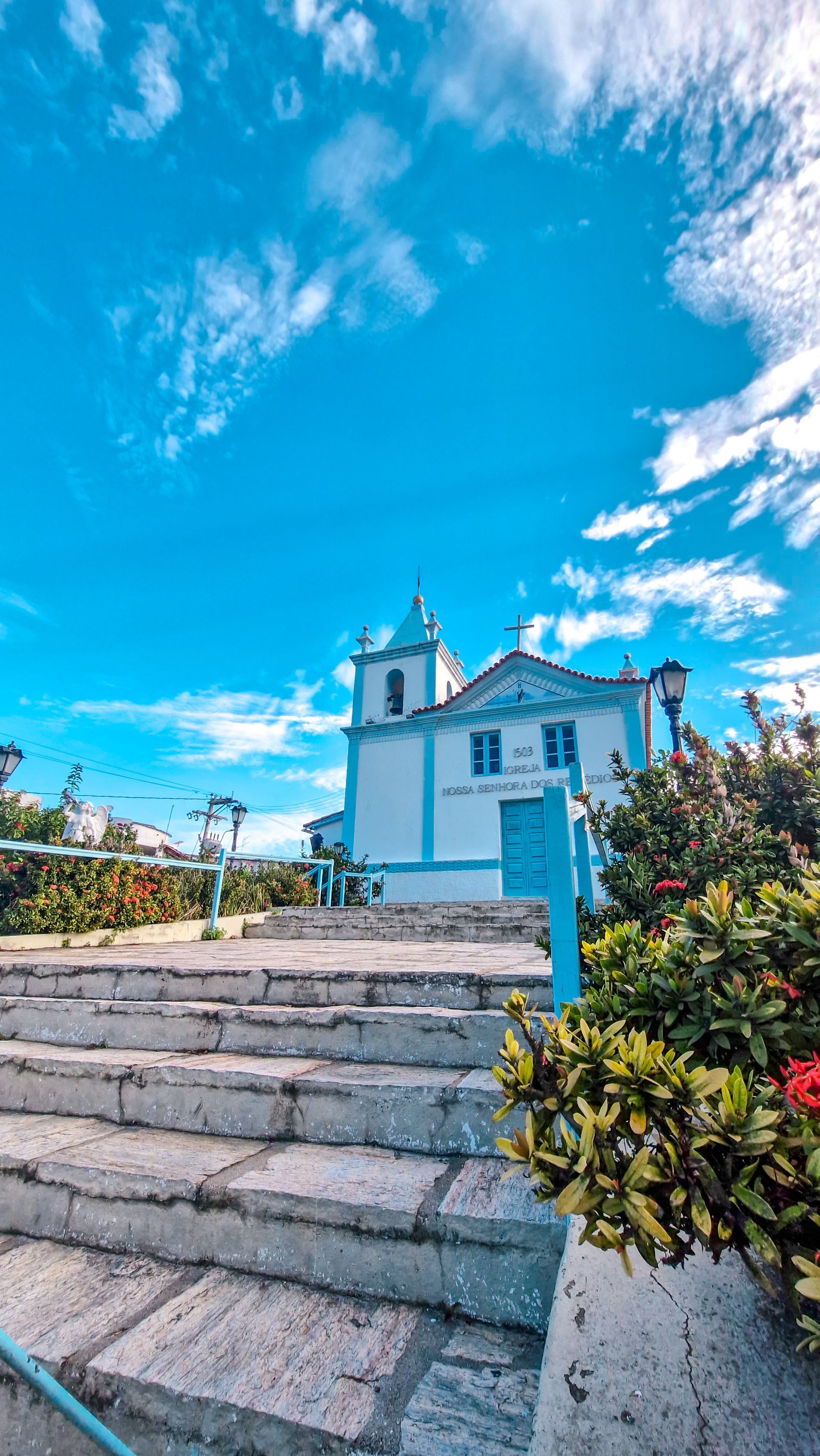 Igreja branca e azul num dia ensolarado, com degraus de pedra que levam até ela, céu azul com nuvens.