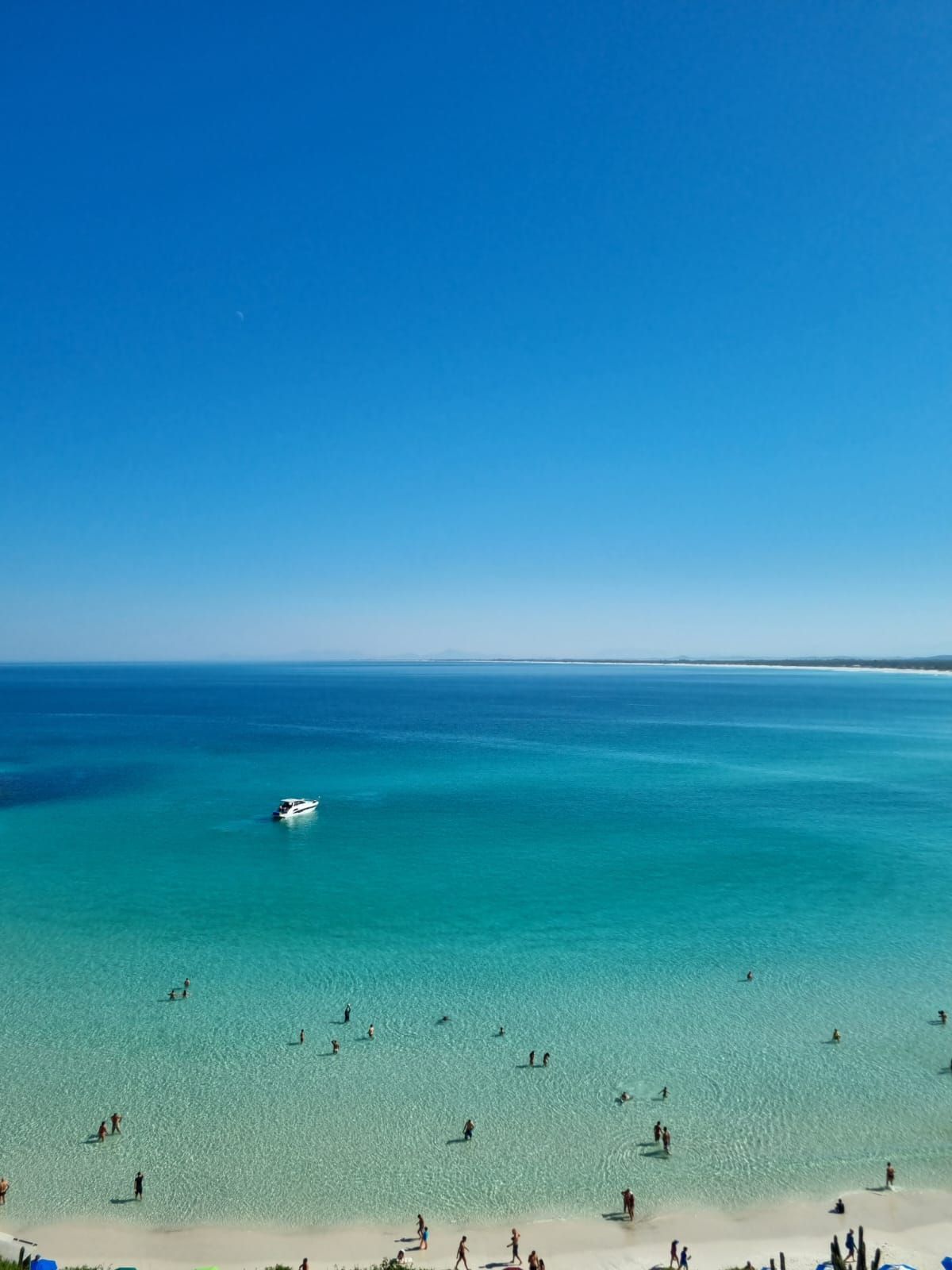 Céu azul brilhante sobre um oceano turquesa com praia de areia branca e pessoas nadando. Um barco é visto ao longe.