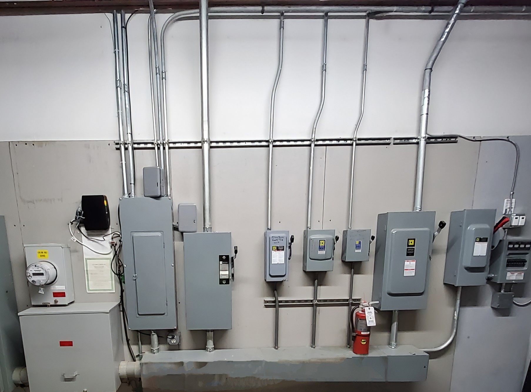 Electrical panel with conduits, meters, and a fire extinguisher against a white wall.