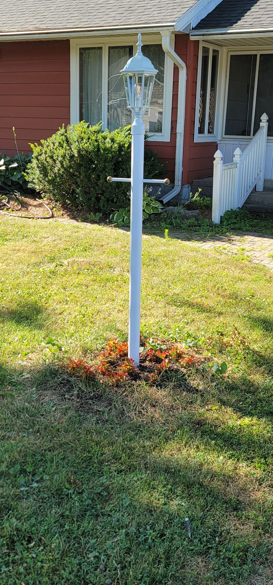 A white bird feeder in a yard with brown patches, in front of a red house.