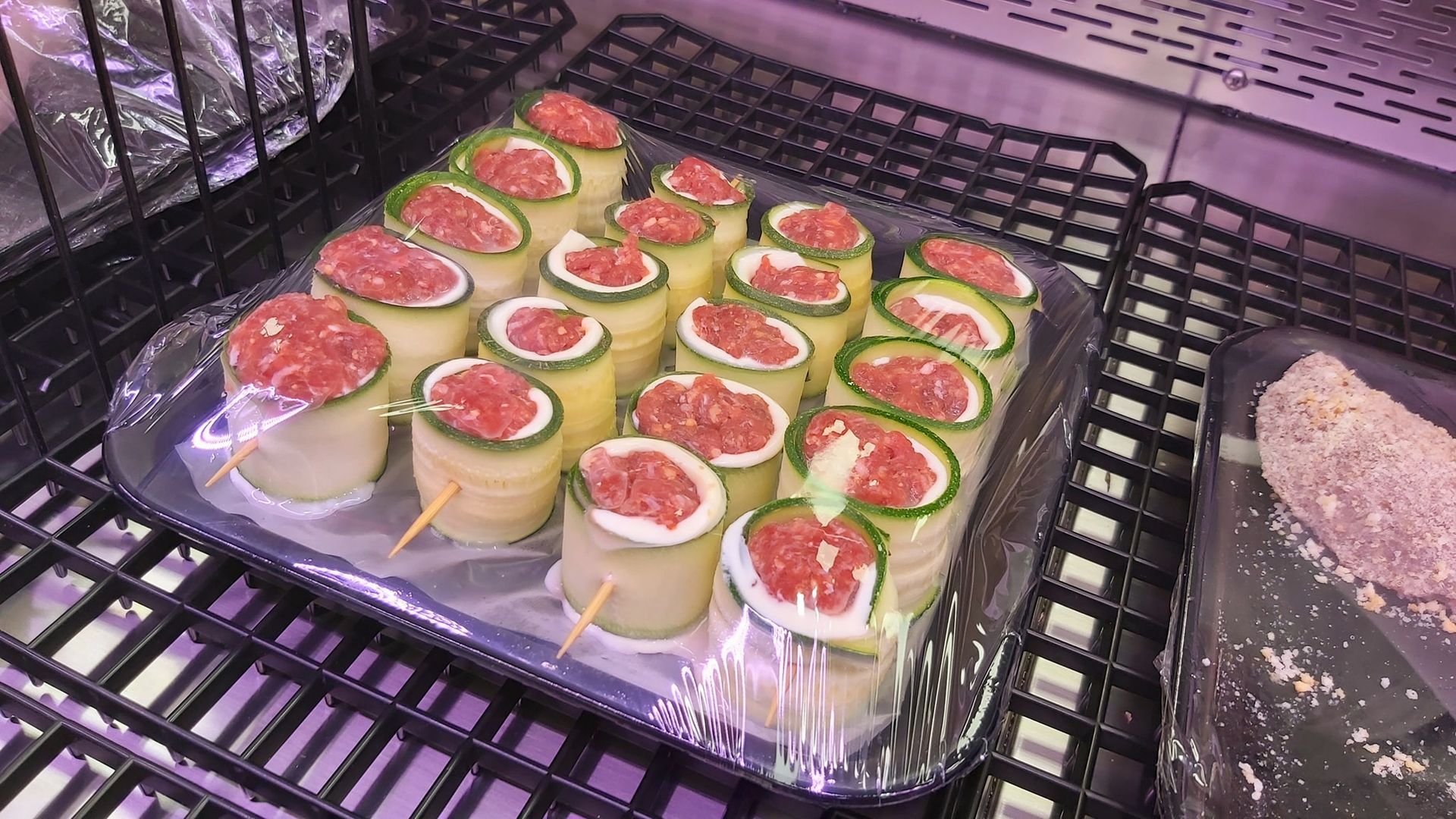 Vassoio di coppette di zucchine affettate con ripieno rosso, adagiate su una griglia da cucina e coperte con pellicola trasparente.