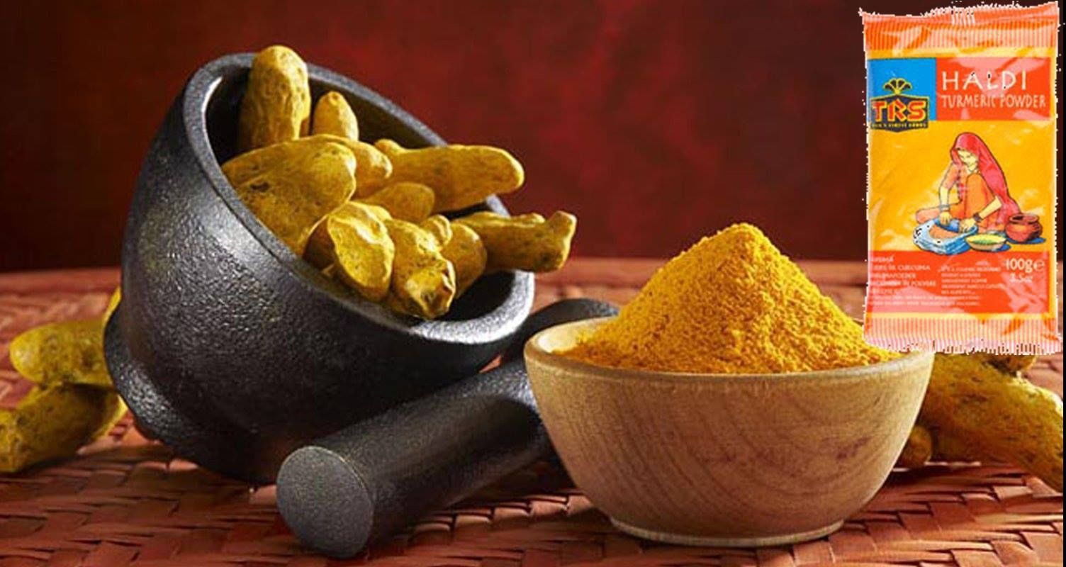 Natura morta rustica con radici e polvere di curcuma in ciotole accanto a una bustina di spezie arancione su sfondo rosso.