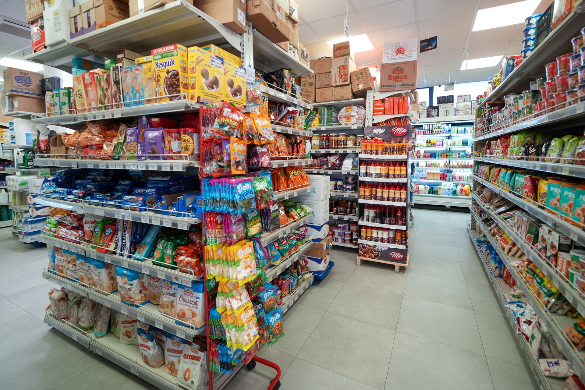 Corridoio di un minimarket con scaffali pieni di snack, caramelle e bevande illuminati da luci fluorescenti intense.