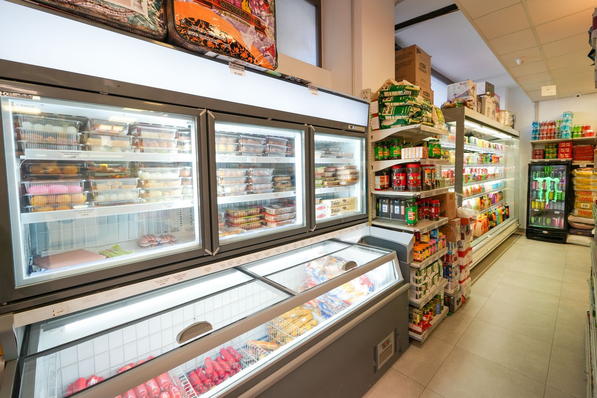 Corridoio del minimarket con bevande refrigerate, snack e scaffali di prodotti confezionati.