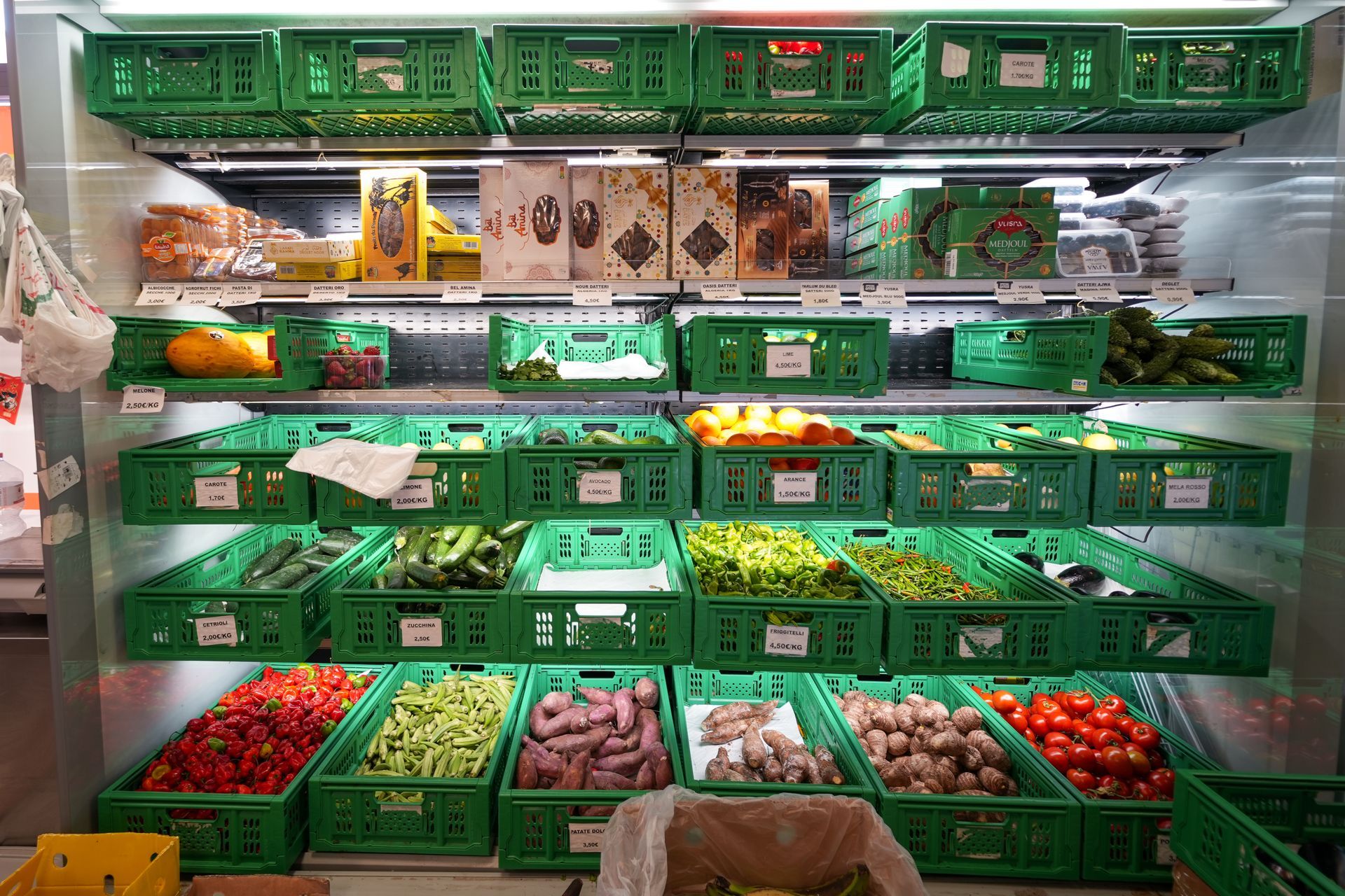 File di cassette verdi piene di frutta e verdura assortita in una vetrina refrigerata.
