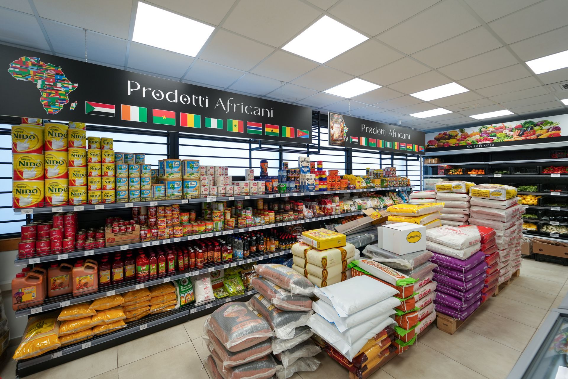 Corridoio ortofrutticolo di un supermercato con pile di sacchi di riso, scaffali di prodotti confezionati e luci intense dall'alto.