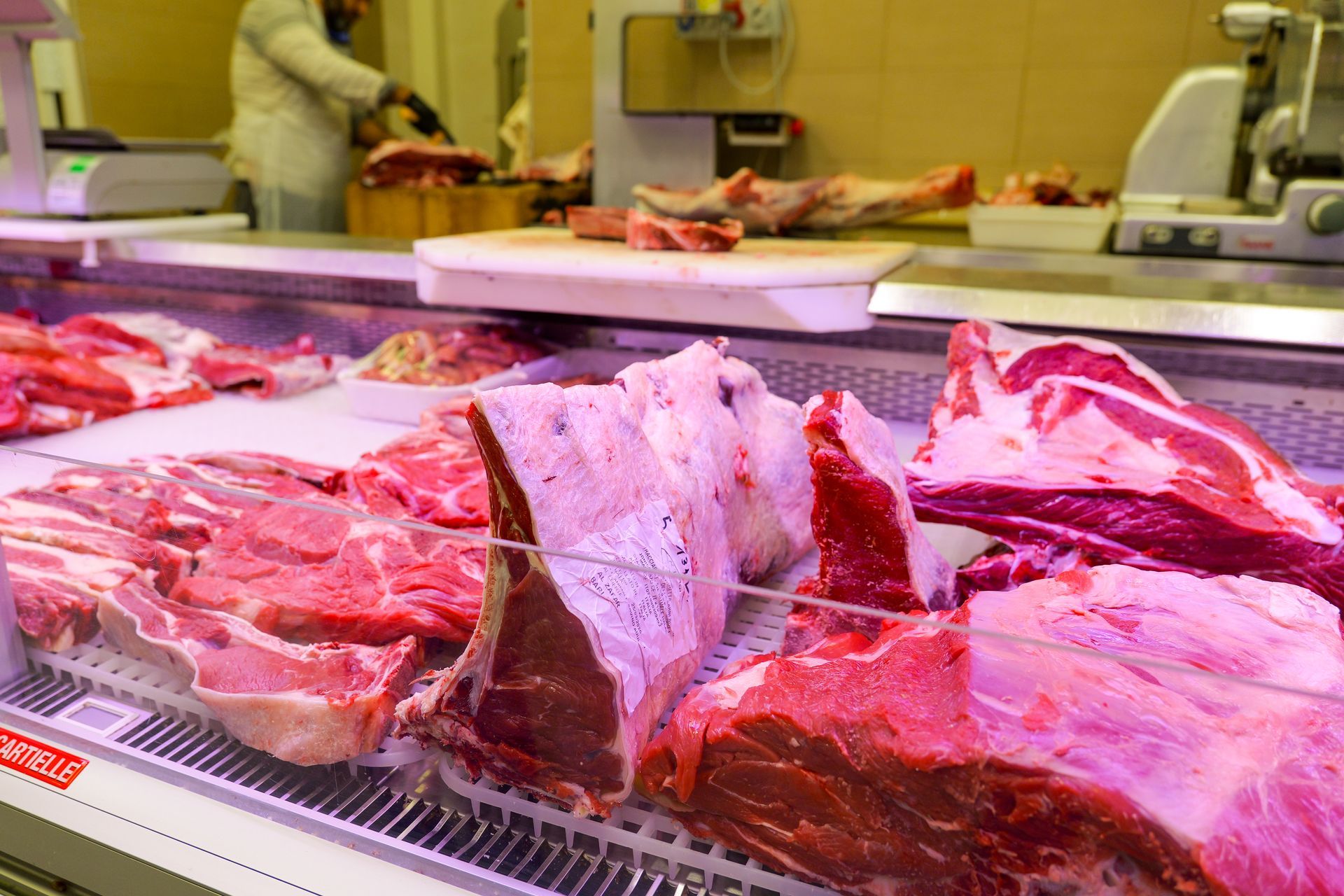 Vetrina di carne in una macelleria con grandi tagli di carne rossa su un bancone e un commesso sullo sfondo.
